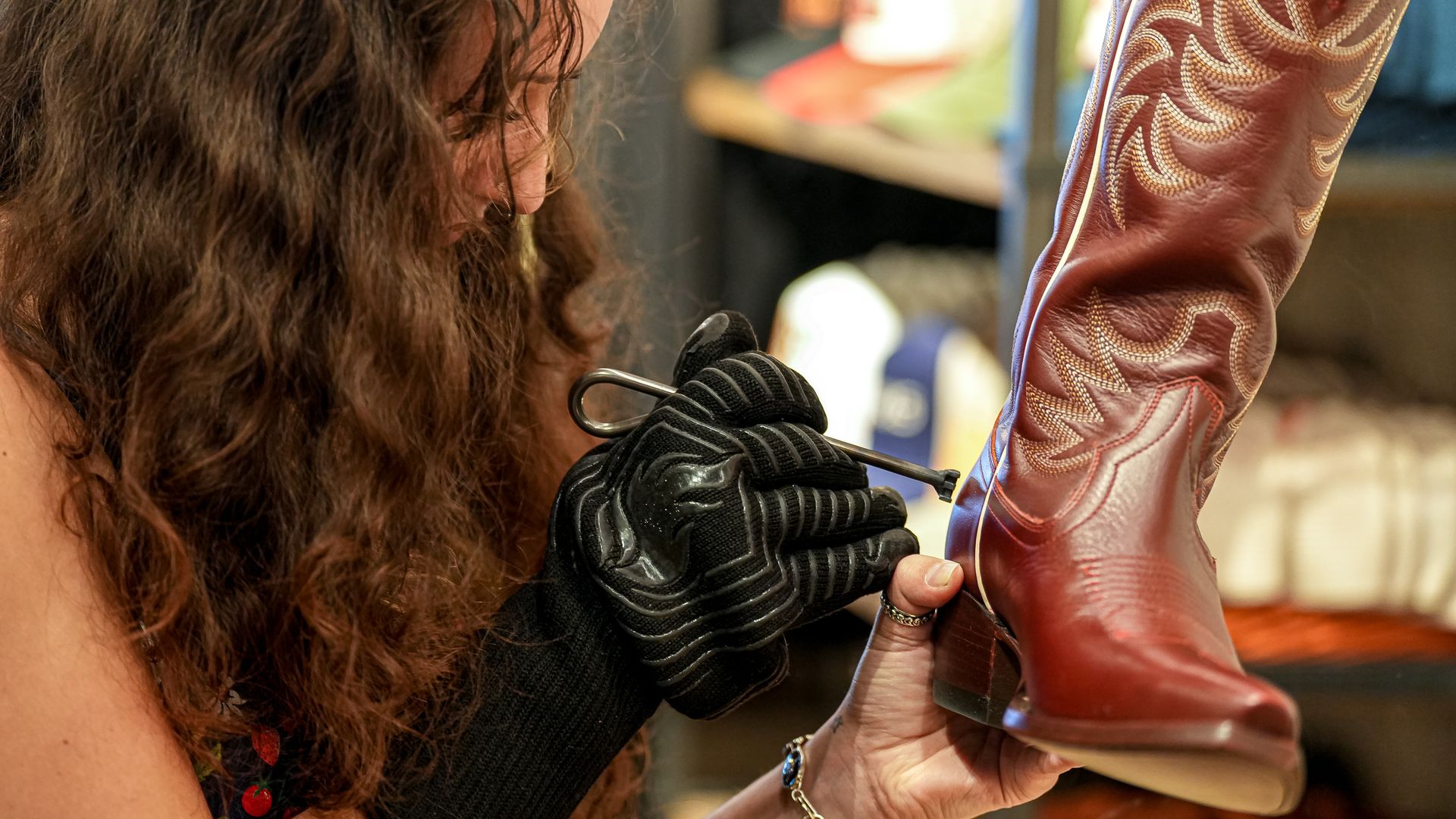 Person with long brown hair wearing a black glove holds a red leather cowboy boot with cream decorative stitching, in a shop setting.
