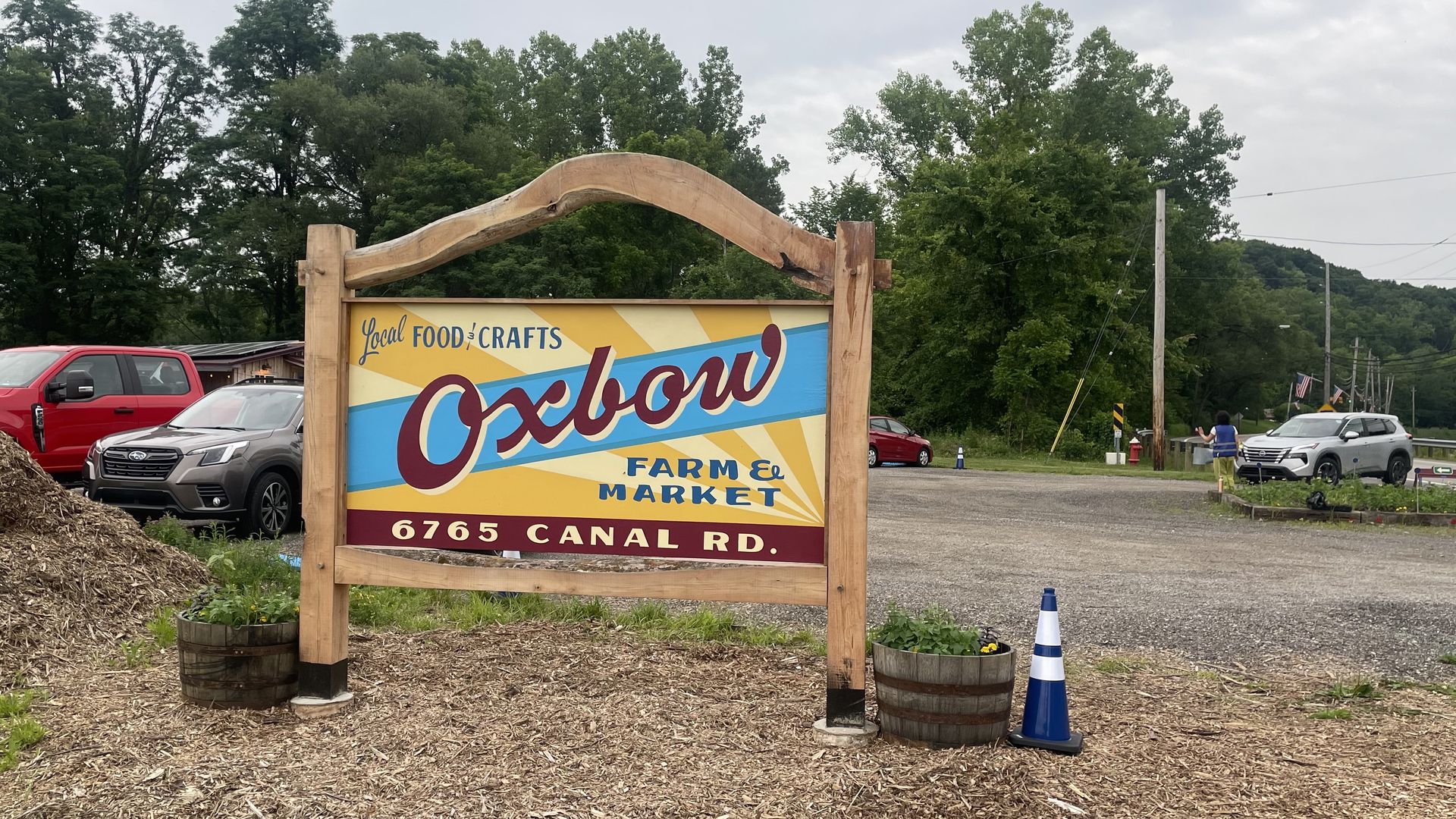 Colorful roadside sign reading "Oxbow Farm & Market" under a curved wooden arch. It says "Local Food & Crafts" on a yellow-blue backdrop; red pickup and gray SUV in a gravel lot, with trees and power lines behind.