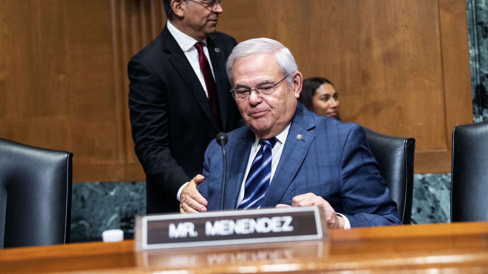 Sen. Bob Menendez, wearing a blue suit, white shirt, blue striped tie and glasses, sits in front of a wooden dais on a black chair.