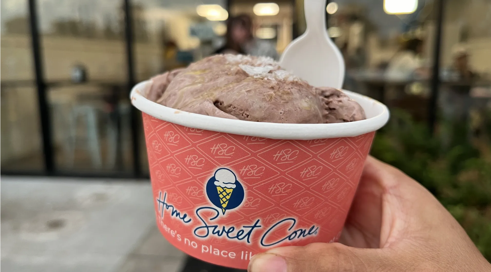 Close-up of a pink ice cream cup with a blue cone logo and the text Home Sweet Cone, holding a scoop of chocolate ice cream with a white plastic spoon, outdoors with a blurred storefront.