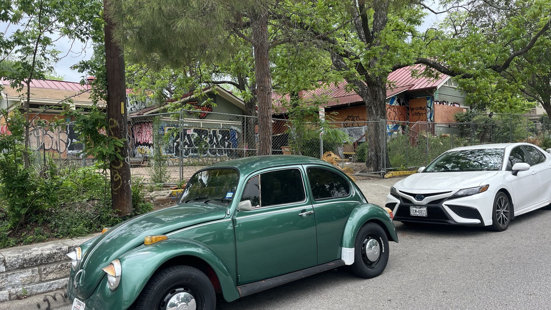 A green vintage Volkswagen Beetle parked on a city street, with a white modern sedan behind it. Graffiti-covered walls, a chain-link fence, and a red-roofed building rise behind leafy trees.