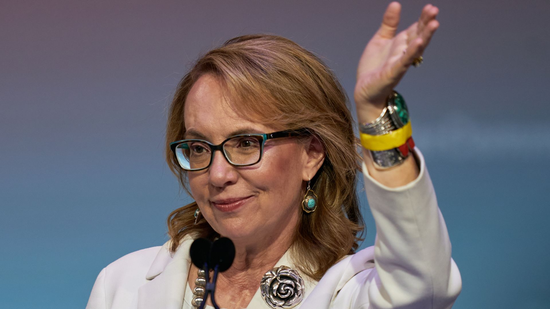 A woman in glasses, wearing a white blazer with a silver-colored flower on the lapel, waves with her left hand. 