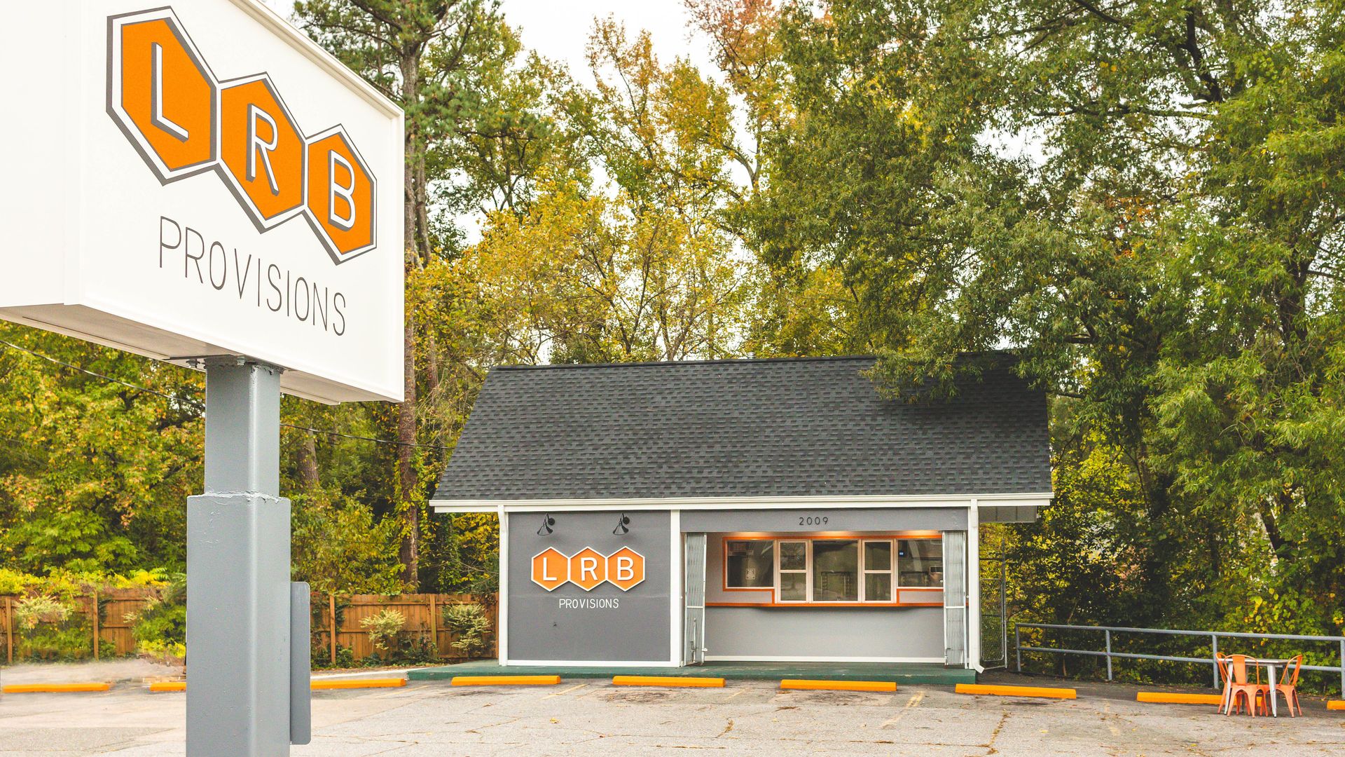 Small gray building with a black roof and orange accents, sign and wall logo read "LRB PROVISIONS," surrounded by trees with autumn leaves and empty parking lot with yellow blocks.