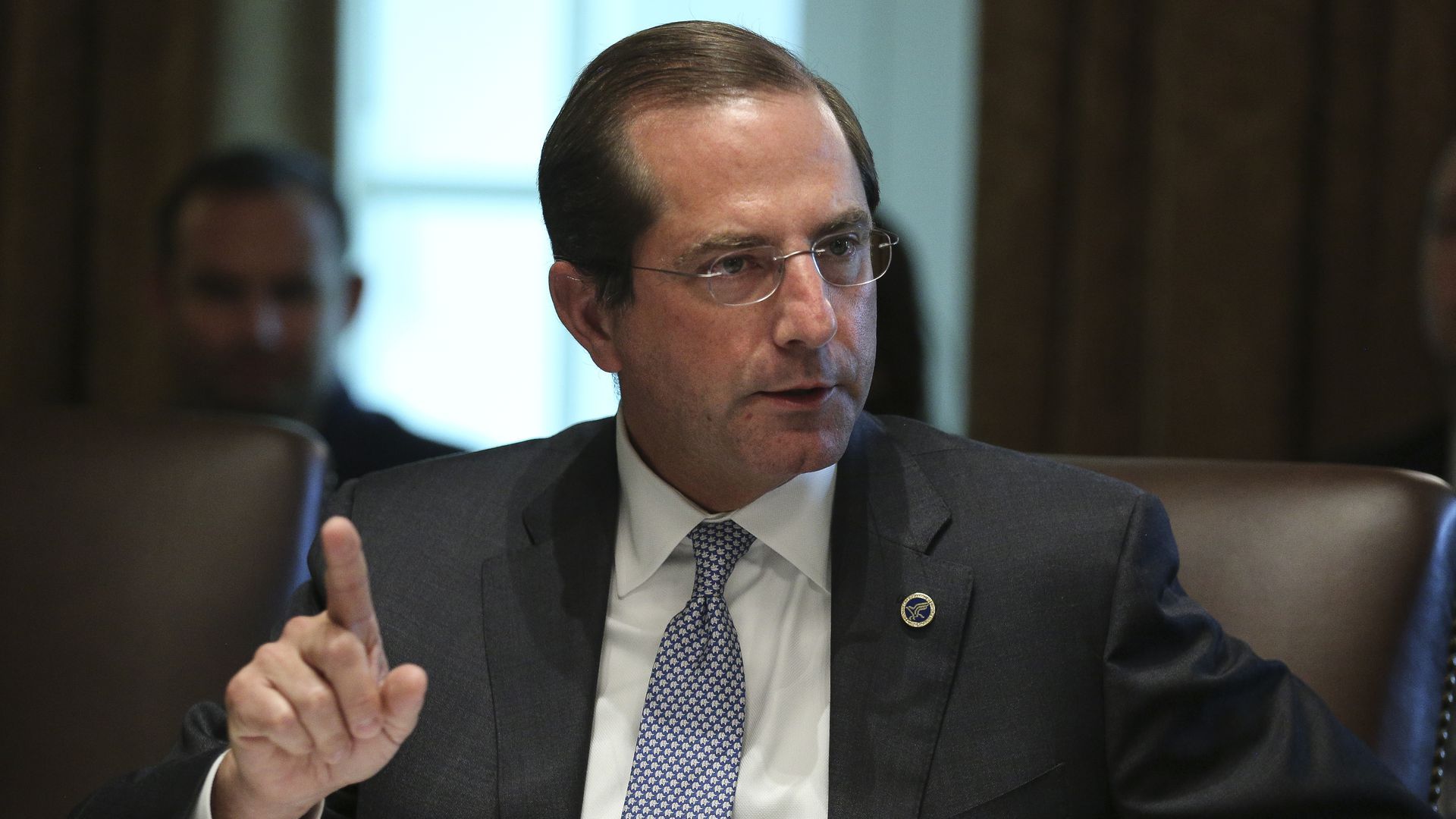 HHS Secretary Alex Azar sits and talks in a brown leather chair, pointing his index finger at the ceiling. 