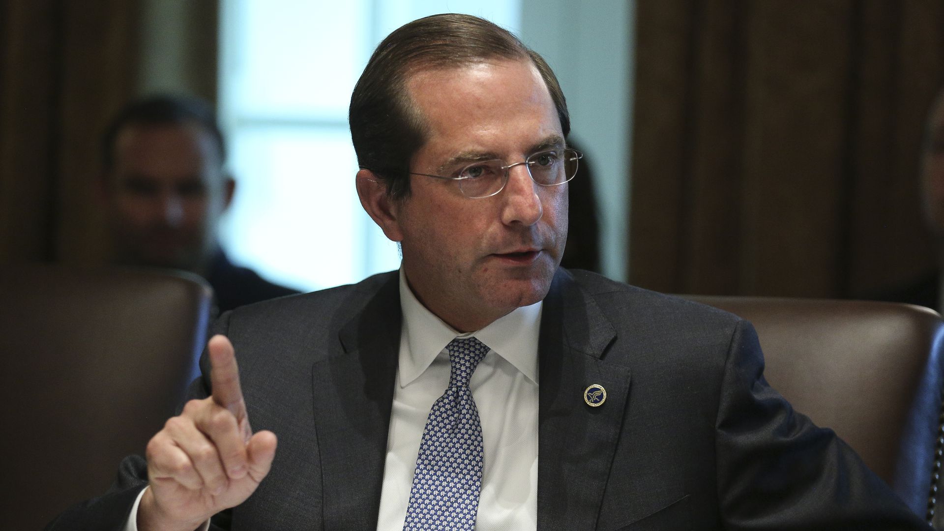 HHS Secretary Alex Azar sits and talks in a brown leather chair, pointing his index finger at the ceiling.