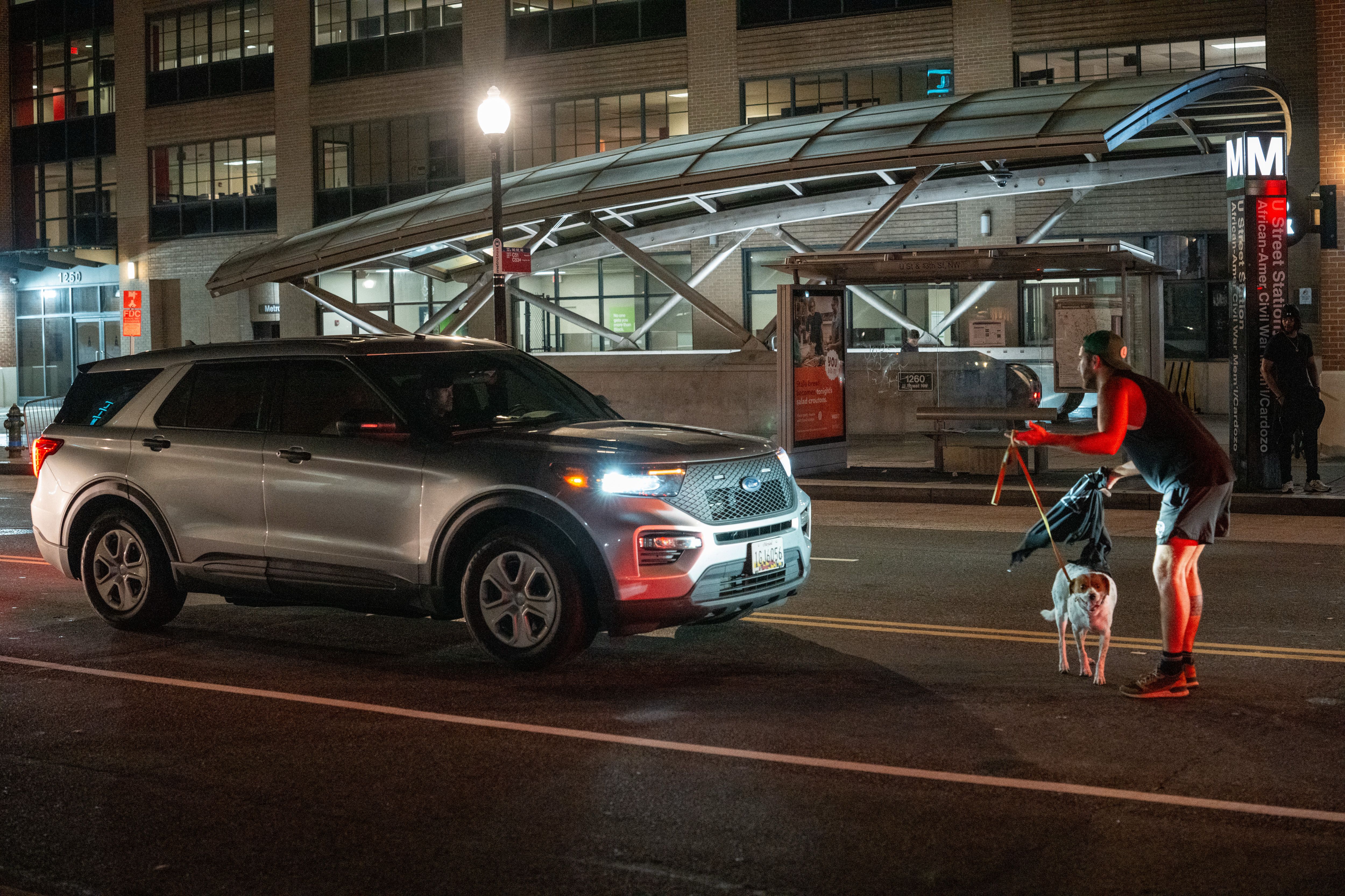 Night scene with a man in shorts and cap holding a dog on leash, standing on the street in front of a silver SUV with headlights on, near a metro station entrance.