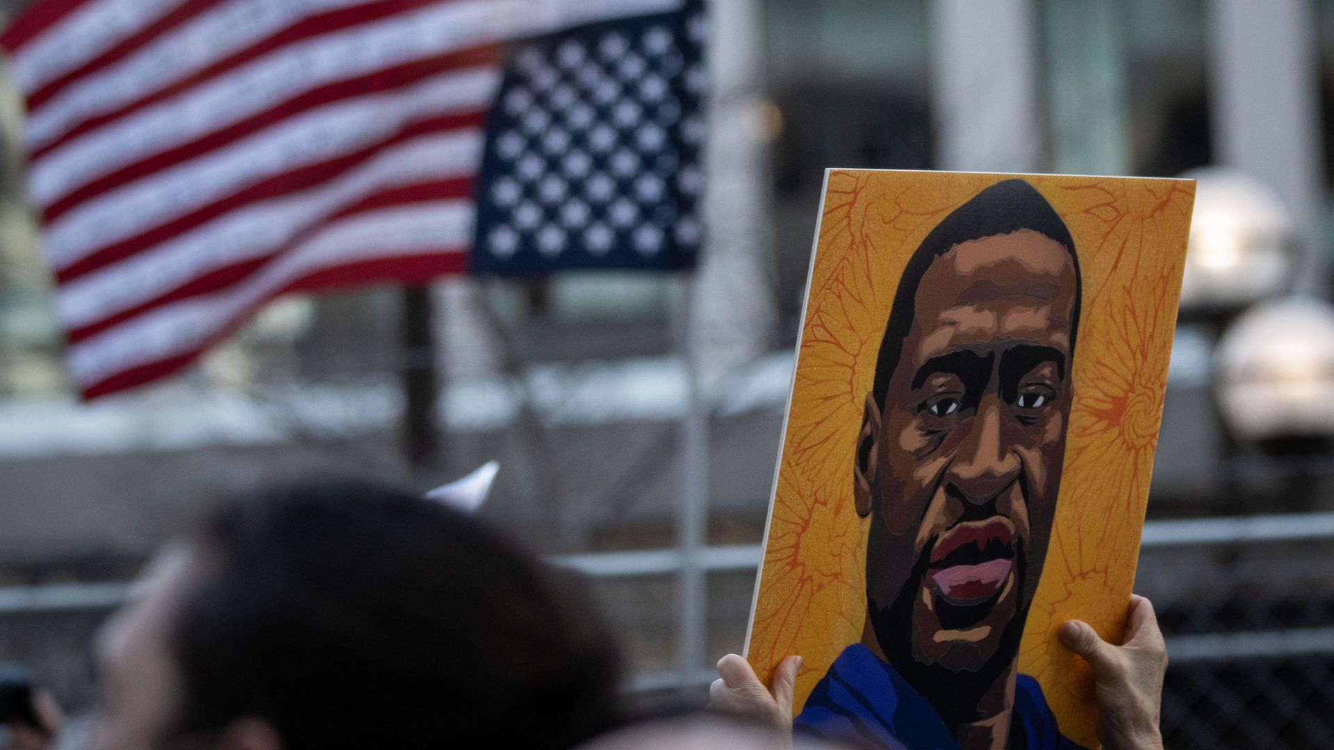 Photo of hands holding up a portrait of George Floyd against an orange background a an American flag flies in the distance