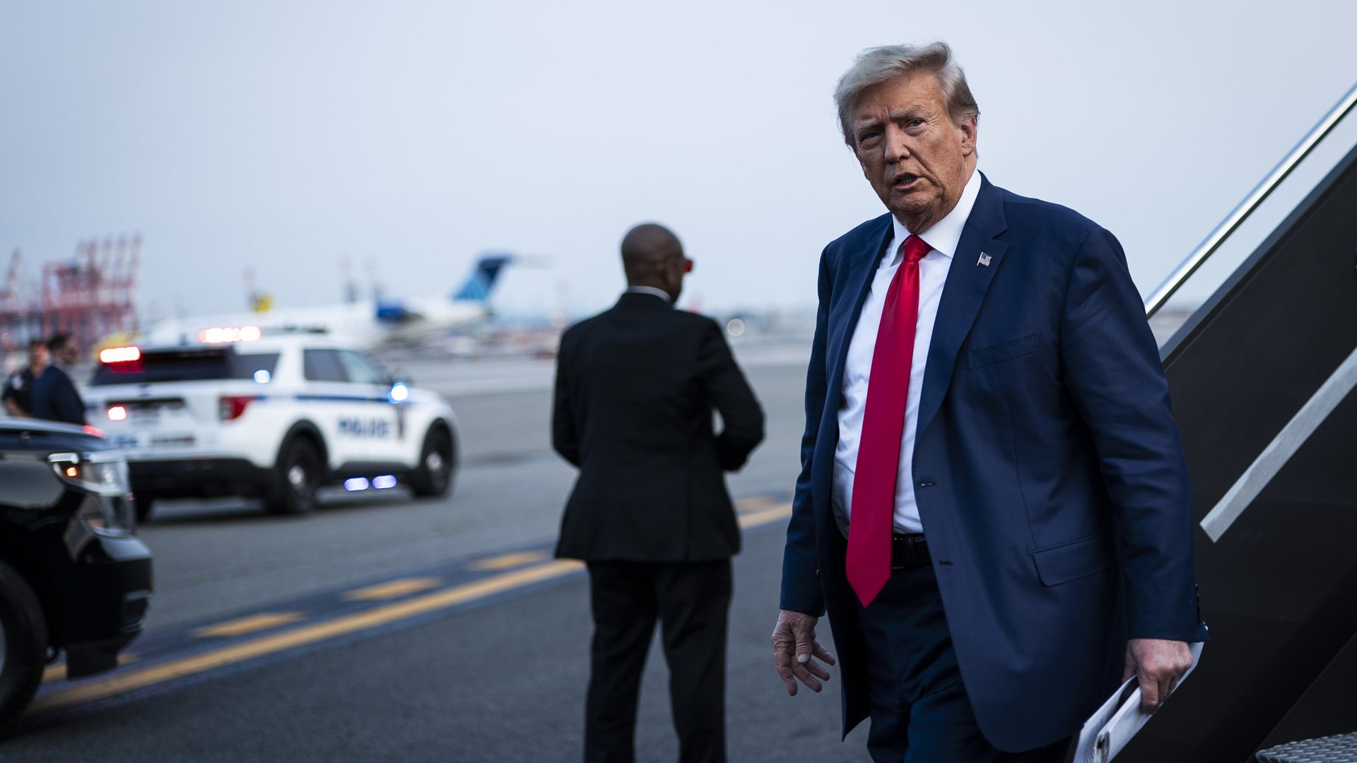 Former President Donald Trump, wearing a blue suit, white shirt and red tie, on an airport tarmac.