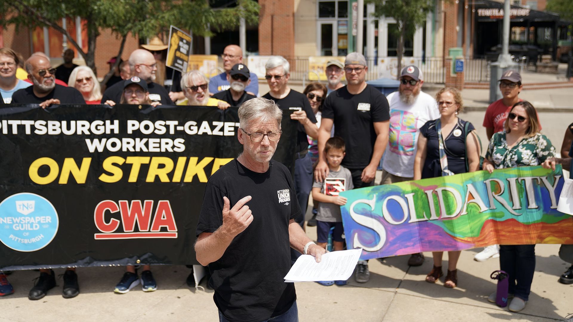 Group of people protesting outdoors with banners reading "Pittsburgh Post-Gazette workers on strike" and colorful "Solidarity" sign, man in front speaking with papers in hand.