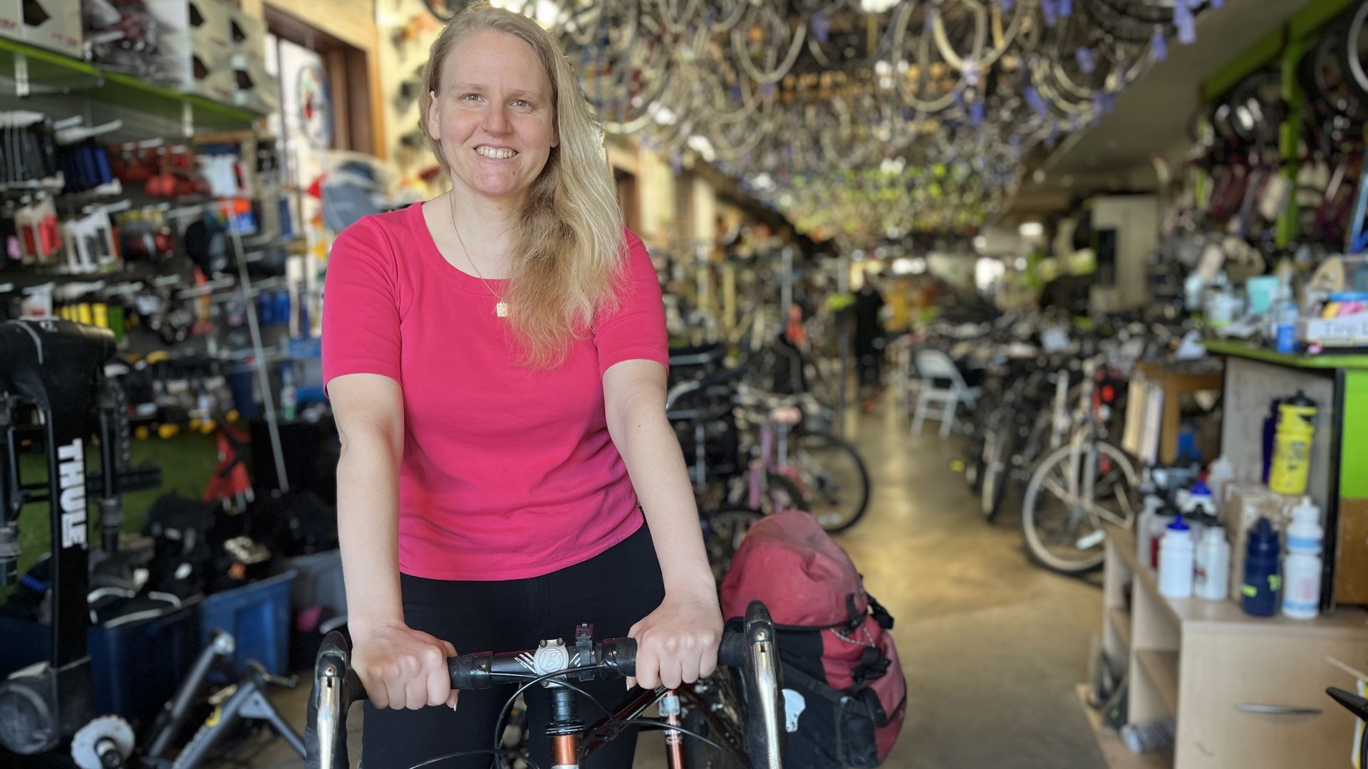 Smiling woman with long blond hair in a pink shirt stands at a bicycle shop, gripping a bike handlebar. The store is lined with shelves of cycling gear and stacked bicycles.