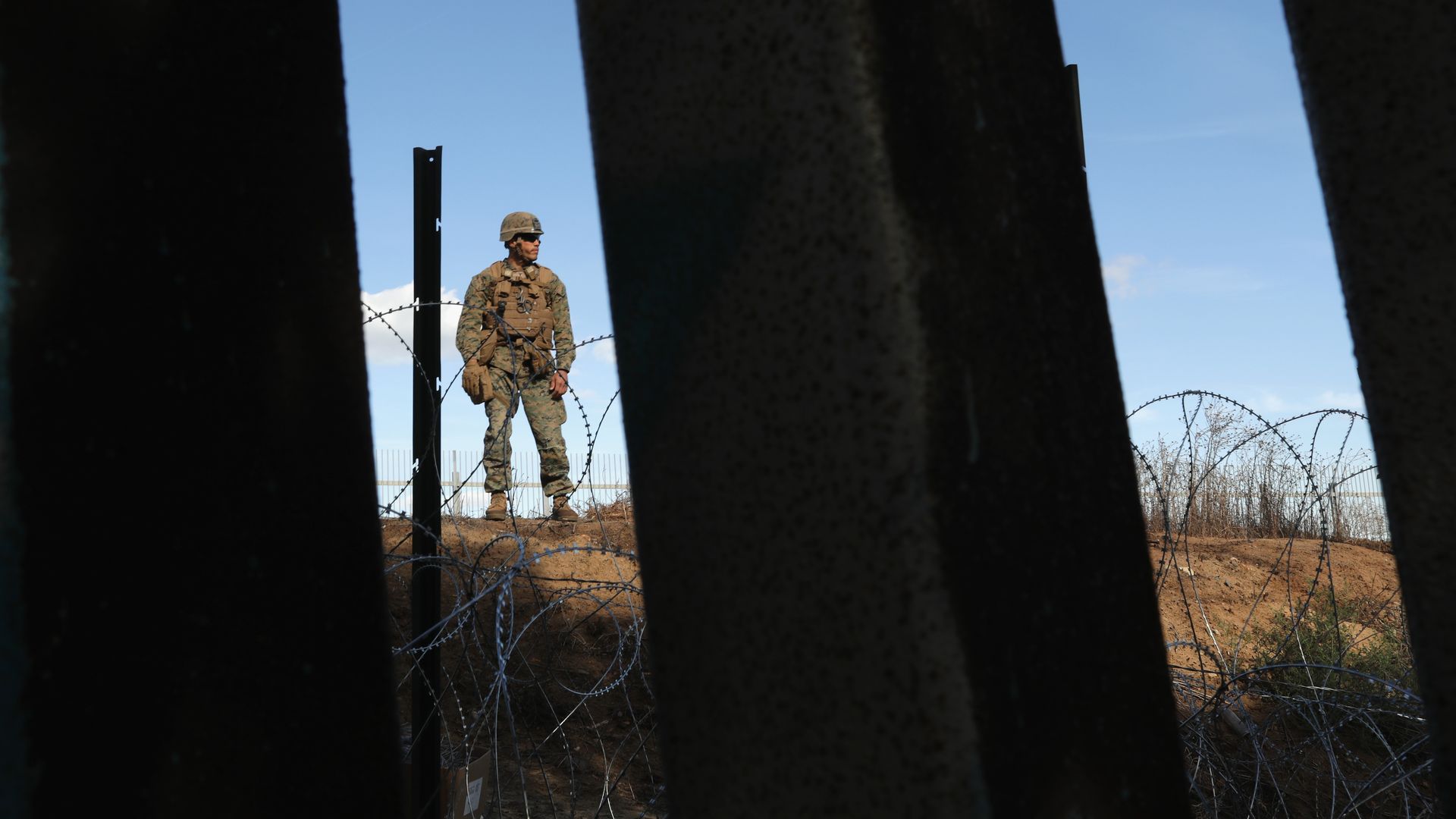 In this image, a Marine is seen in the distance between two fence poles that are close up against the camera. The clear sky is behind the Marine, who is in full uniform.