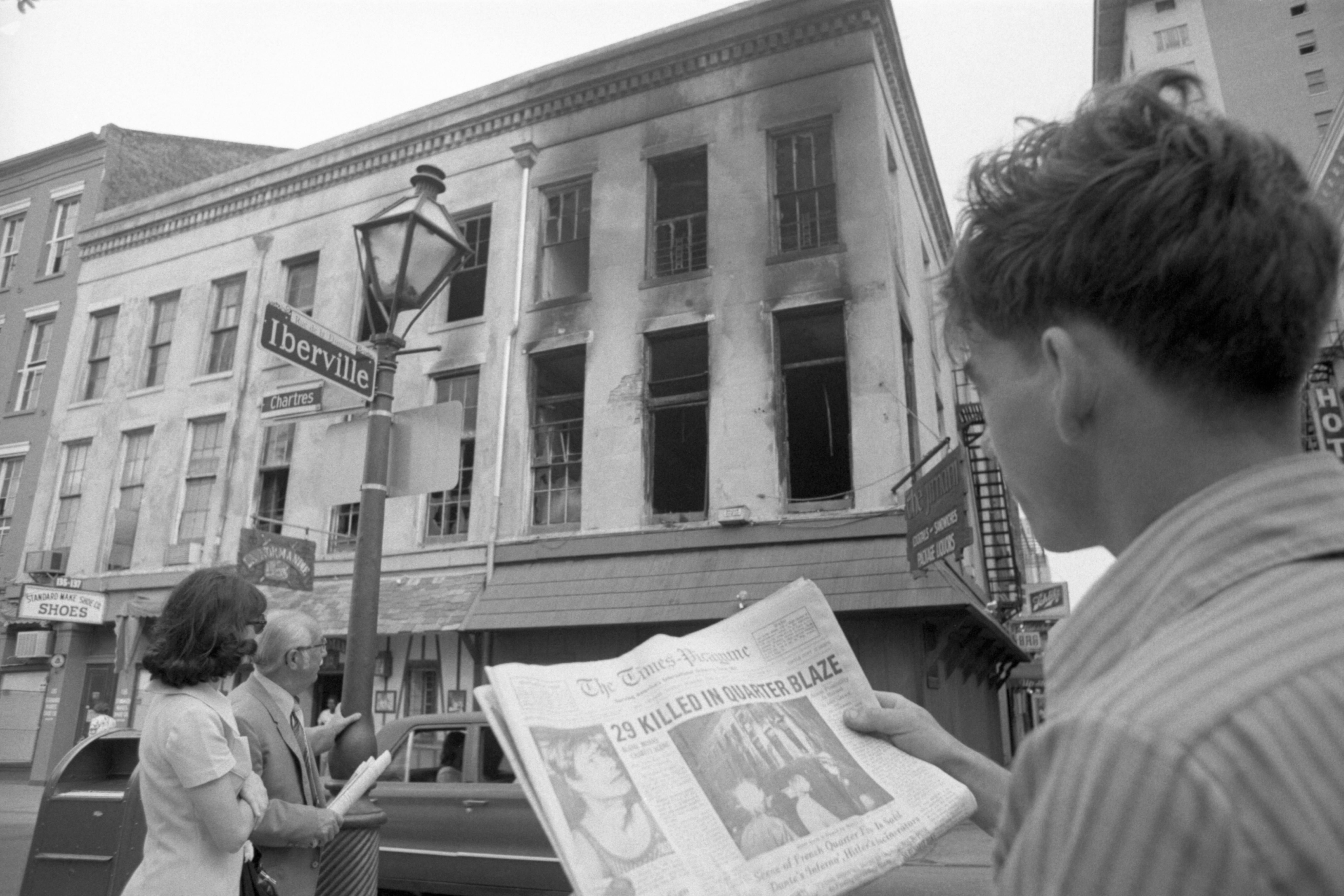 Image shows a man holding a newspaper that says "29 killed in Quarter blaze" in front of the burned Up Stairs Lounge building.