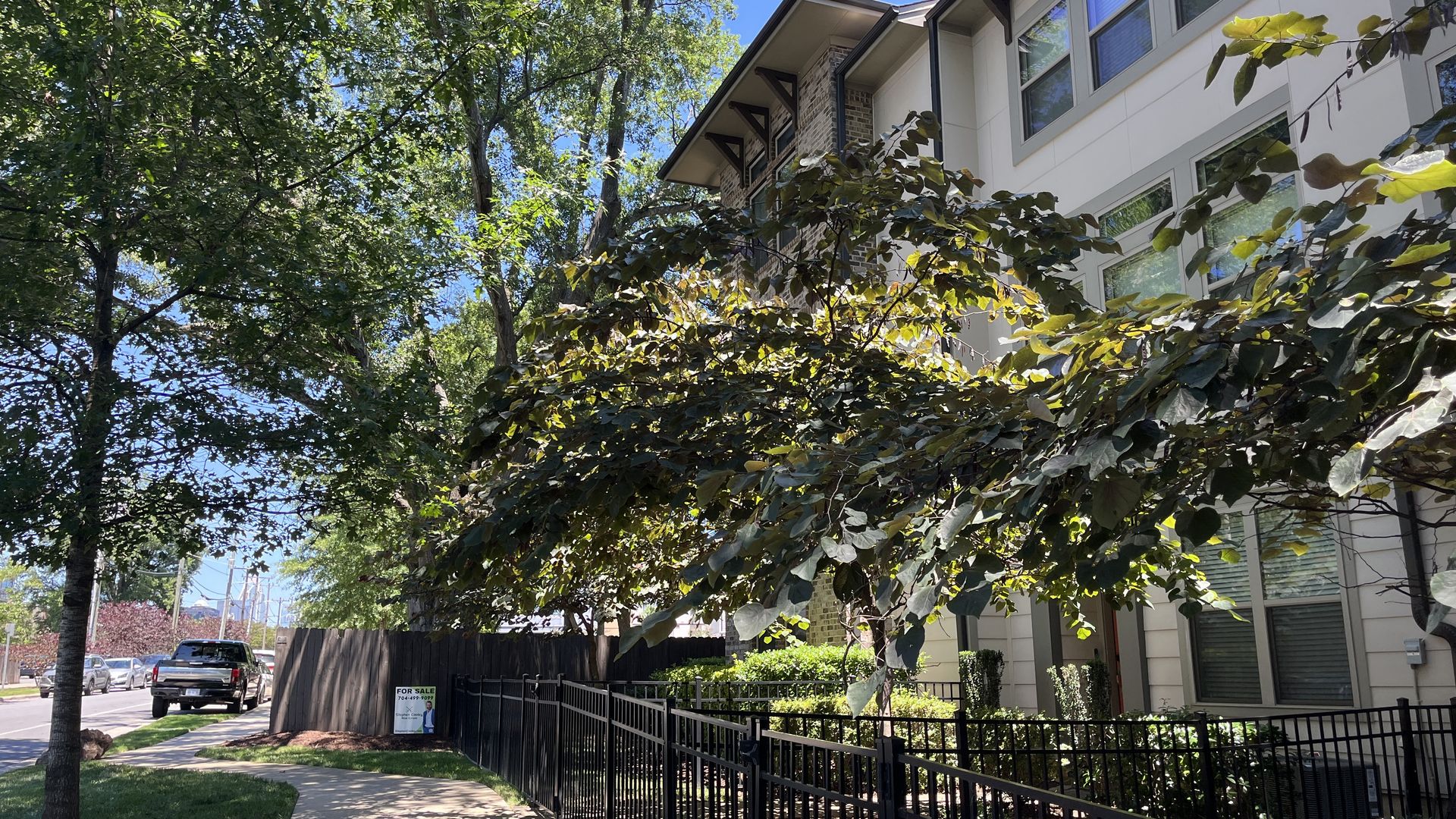 Sunny sidewalk beside a modern beige building with large windows, black metal fence, and overhanging tree branches. A "FOR SALE" sign on the fence; parked cars along the street, blue sky.