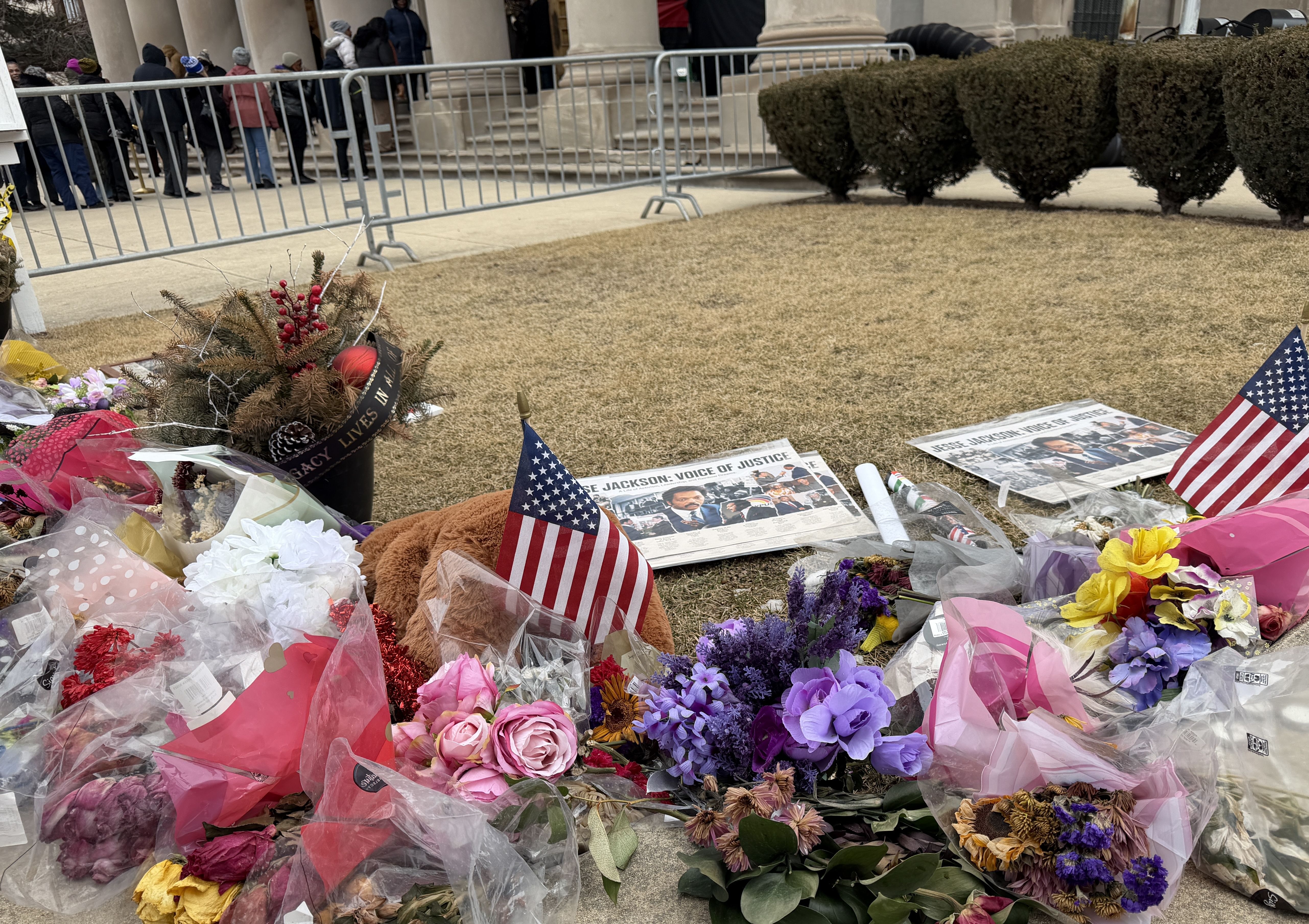 Memorial with colorful flowers, American flags, and signs reading "Jesse Jackson: Voice of Justice" placed on grass outside a building with columns and a line of people.