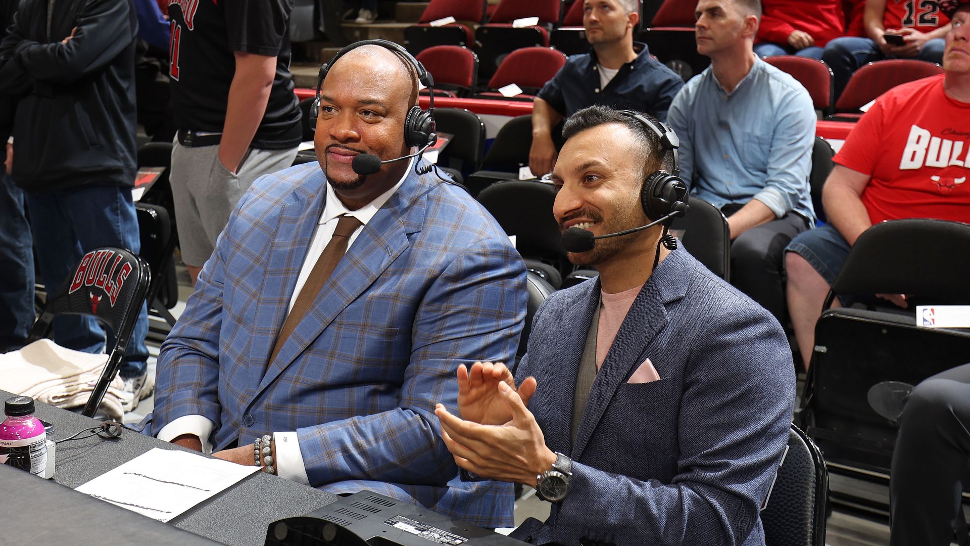 Two men in suits wearing headsets sit at a broadcast table in a basketball arena, microphones in front and papers on the table. Red arena seats and fans in Bulls gear visible in the background.