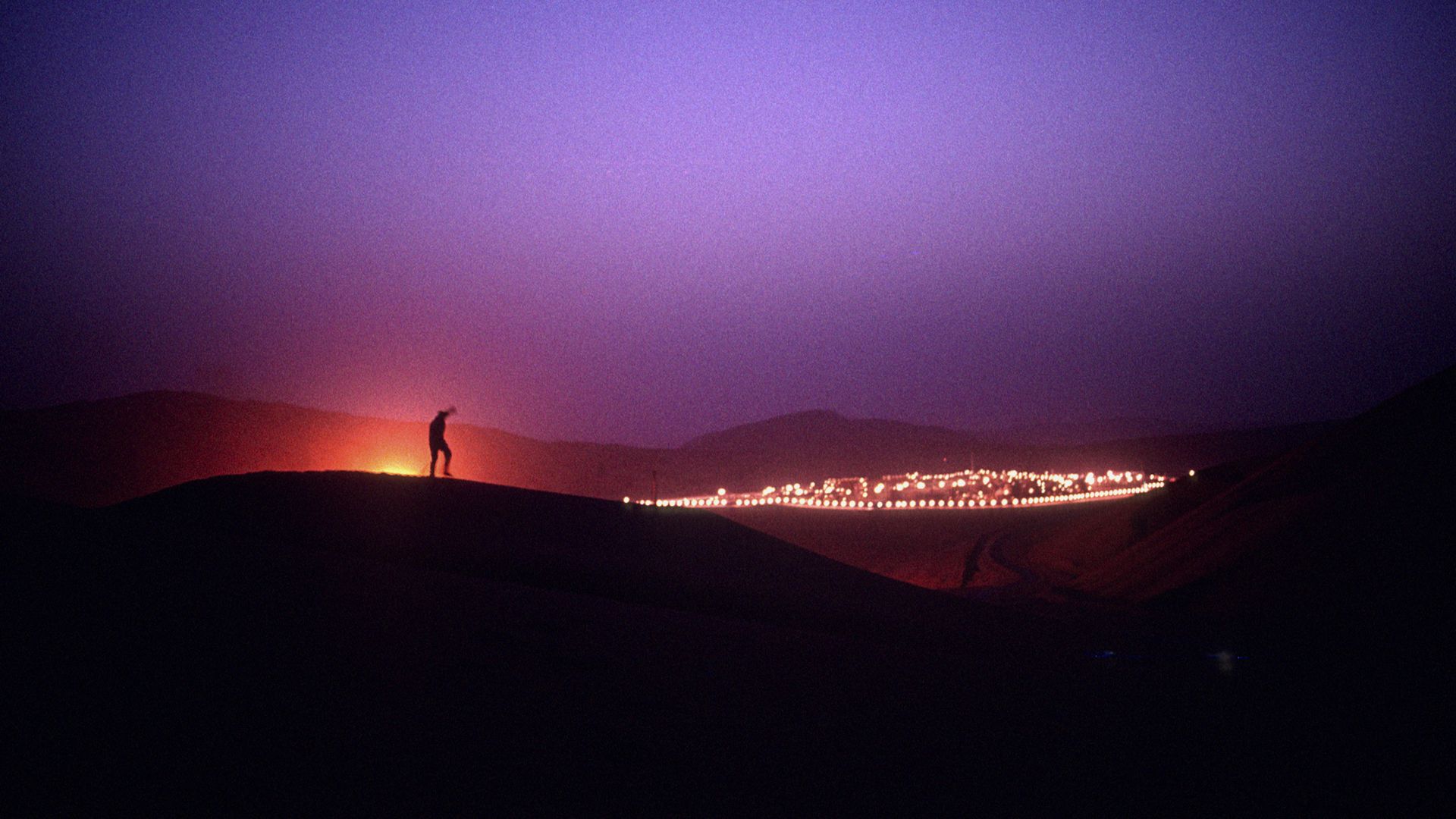 A man stands on a sand dune at night outside of a Saudi Aramco oil field complex