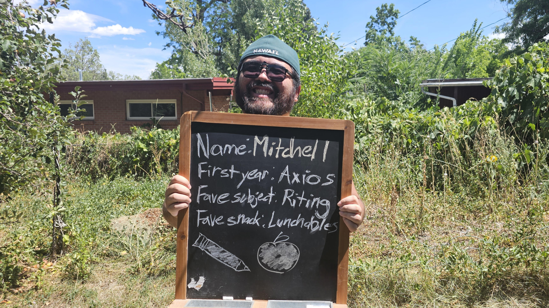 Smiling man wearing a green hat and sunglasses holds a chalkboard outdoors with text about himself: Name Mitchell, First year Axios, Fave subject Writing, Fave snack Lunchables.