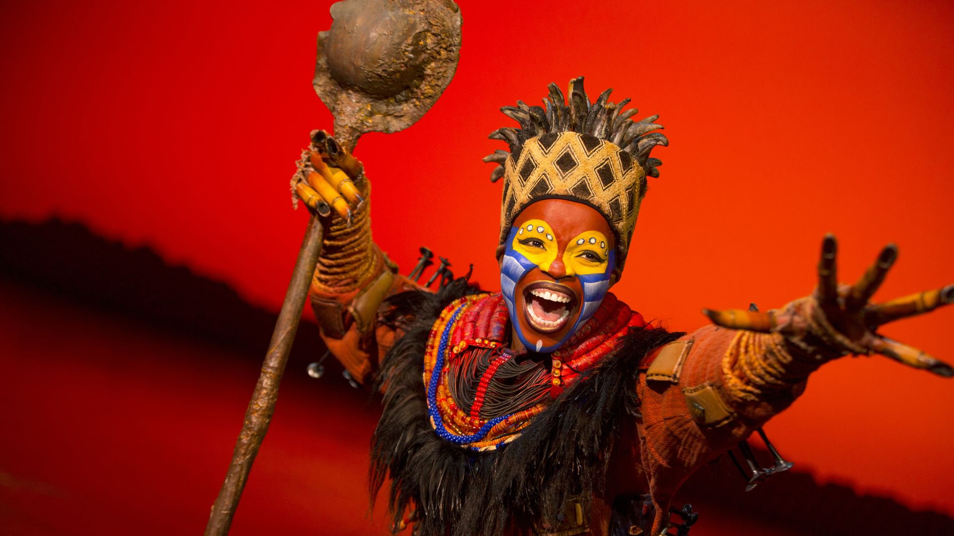 Smiling performer with red, yellow, and blue face paint wears a crown and colorful beads, holding a staff against a vivid red background in a theatrical costume.