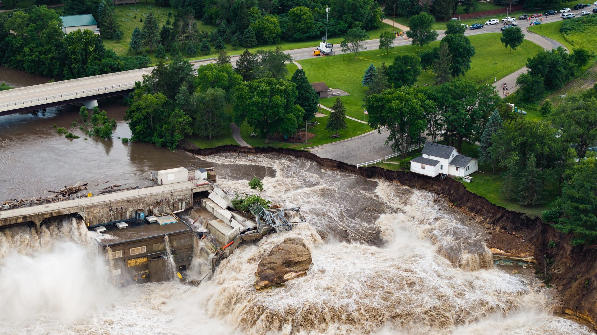 An aerial image of a dam failure in progress, showing silty brownish waters of a rushing river rushing around an abutment of a concrete structure, eroding through a roadway, and rushing forcefully downstream.
