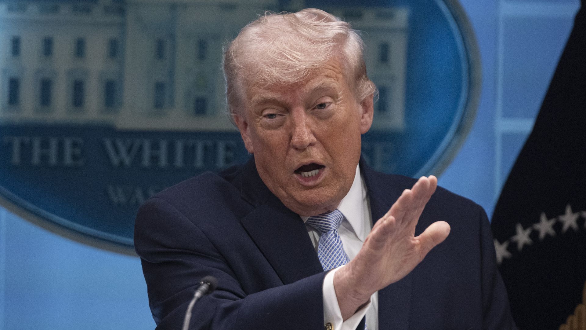 Close-up of a man with light hair in a dark suit and blue striped tie speaking at a press event, gesturing with his hands; a blue backdrop with a round emblem behind him.