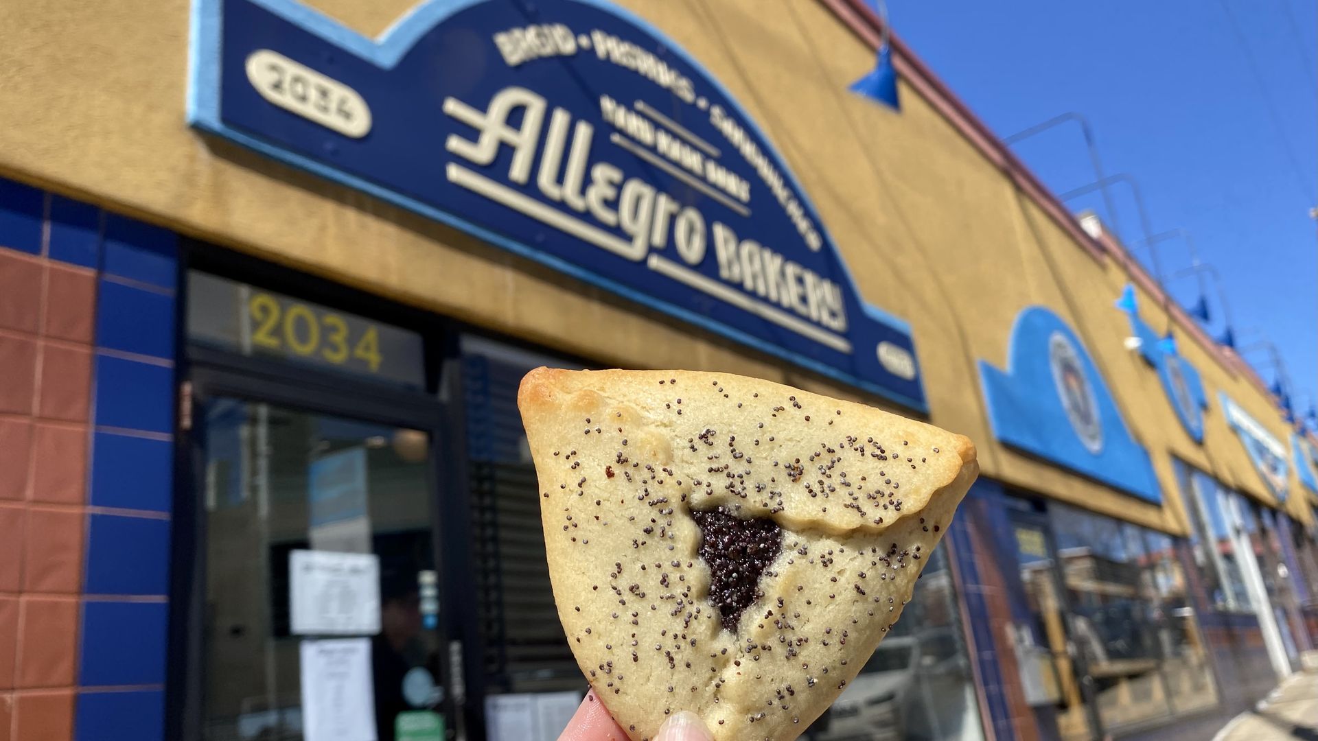 Hand holding a triangular poppy seed pastry in front of Allegro Bakery with blue and yellow signage on a sunny day at 2034 street address.