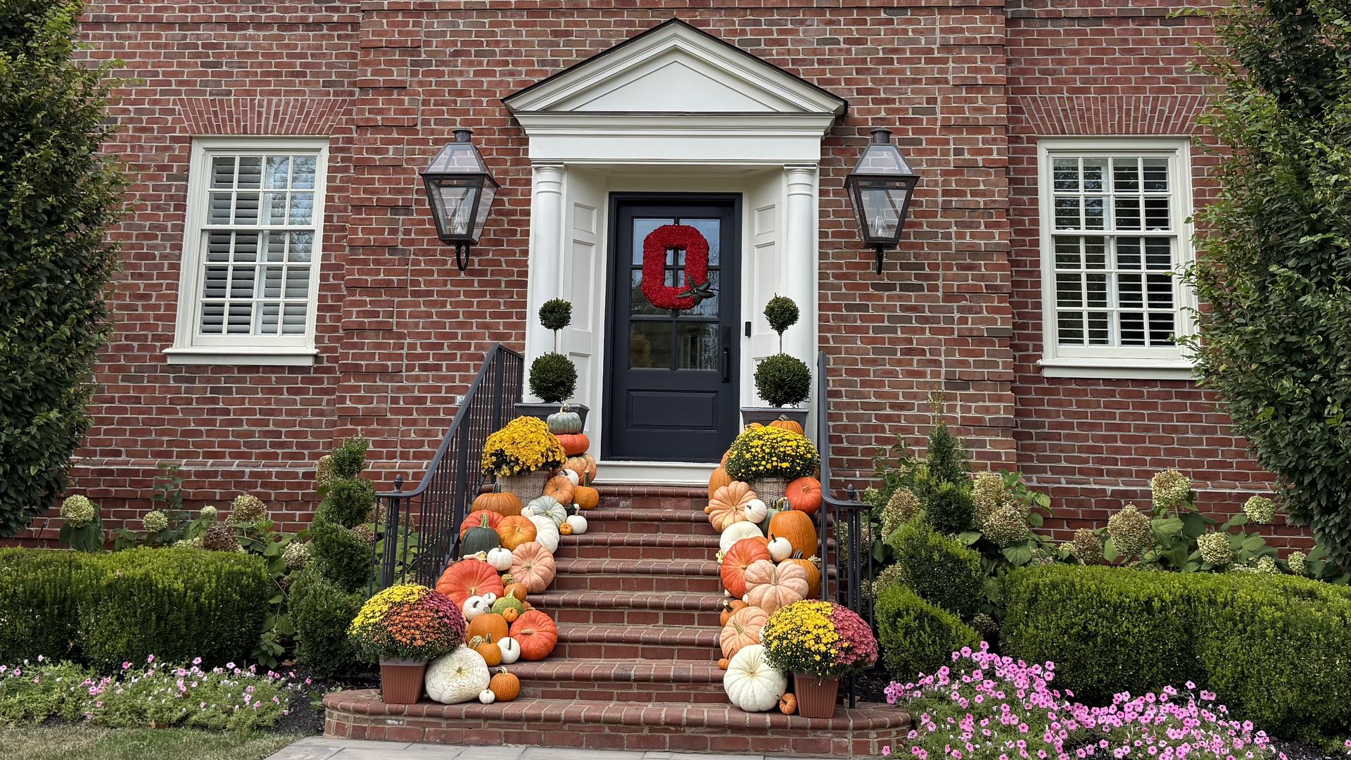 Two-story red brick house with white-trimmed windows and steps lined with orange, white pumpkins and yellow mums.