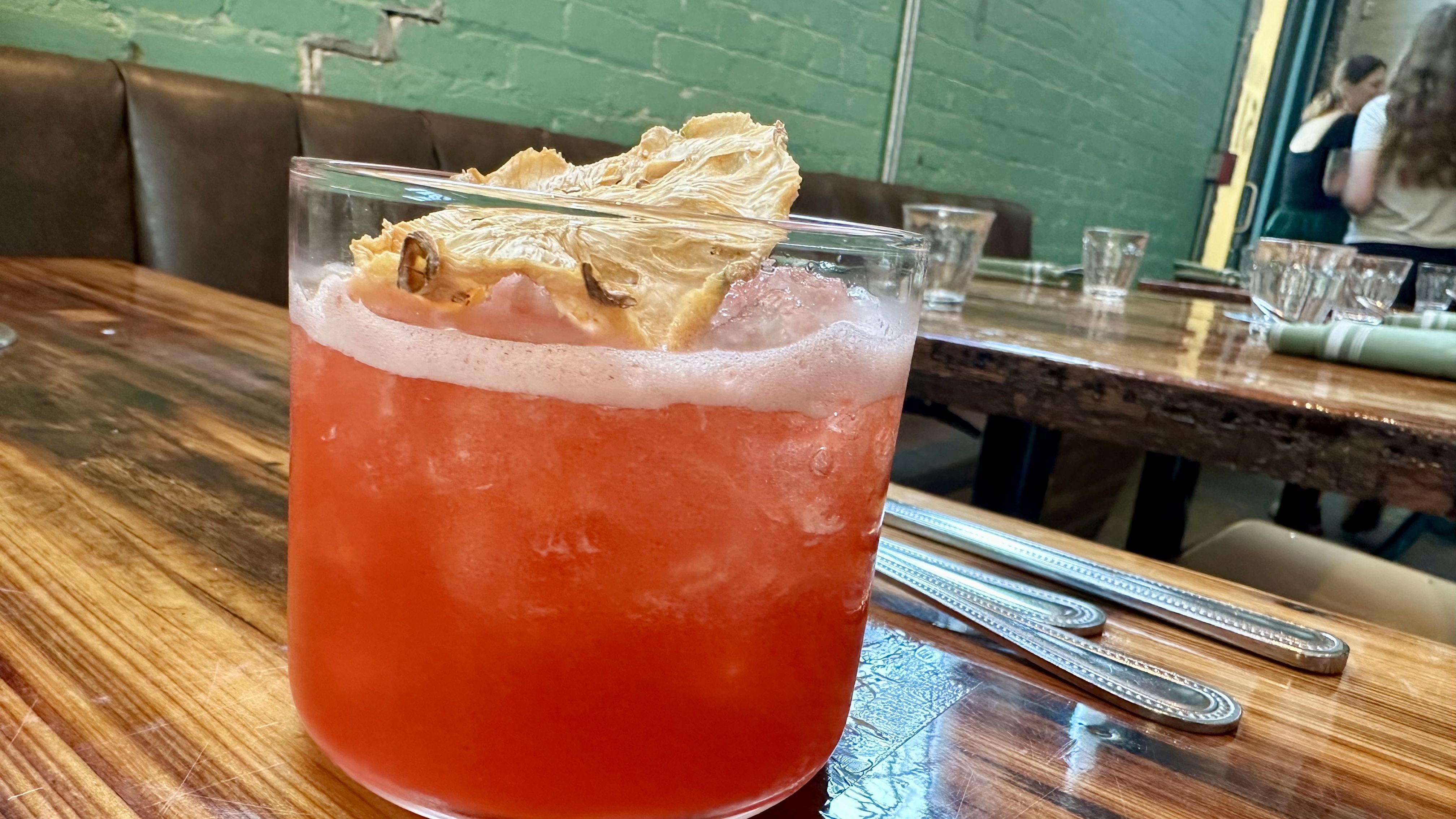 Close-up of a bright red cocktail in a clear glass with a large dried pineapple slice garnish, foamy edge, on a rustic wooden bar table with a green brick wall in the background.