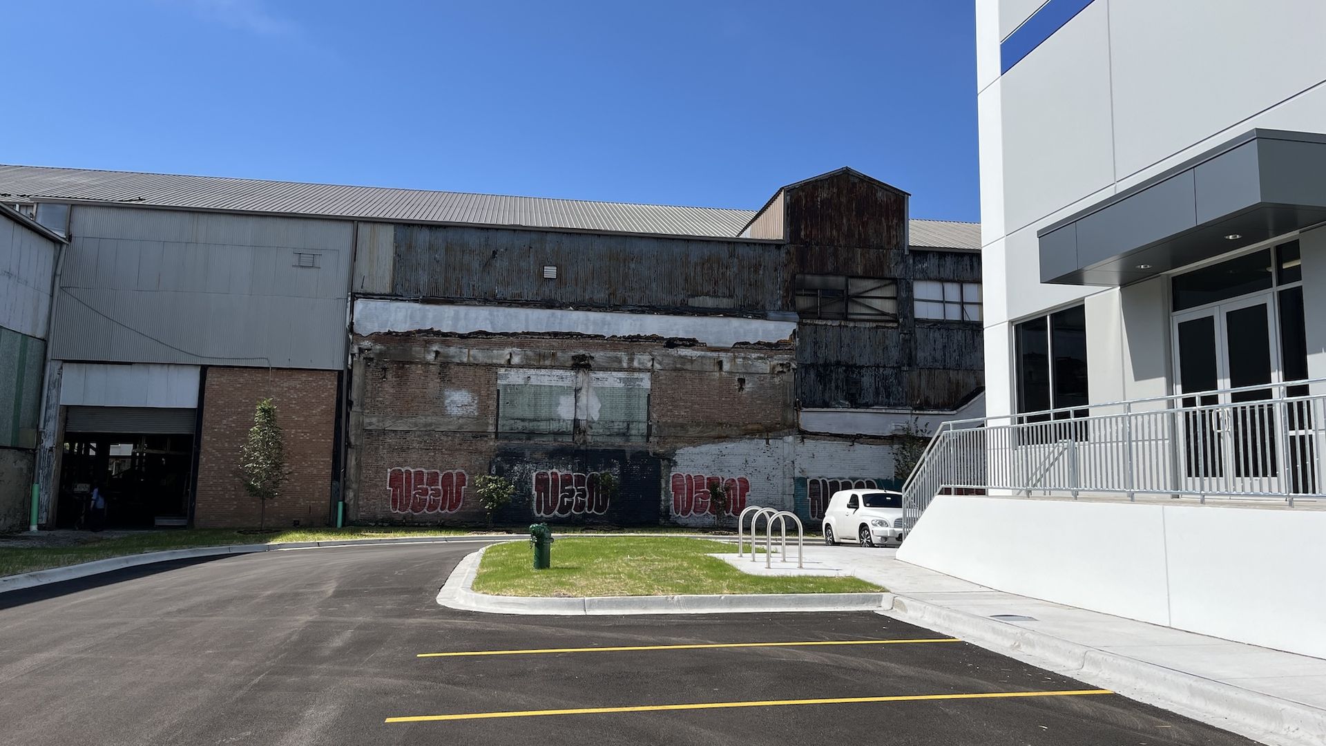 Blue sky over a modern white building with a blue stripe on the right; a rusted warehouse sits left. Graffiti on a brick wall, a white car, a green fire hydrant, and a curved road in the foreground.