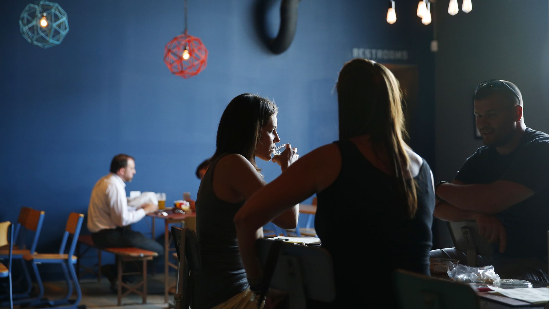 People sitting and talking in a dimly lit cafe with blue walls, hanging geometric pendant lights, and exposed pipes. One woman drinks from a glass while others chat around wooden tables.