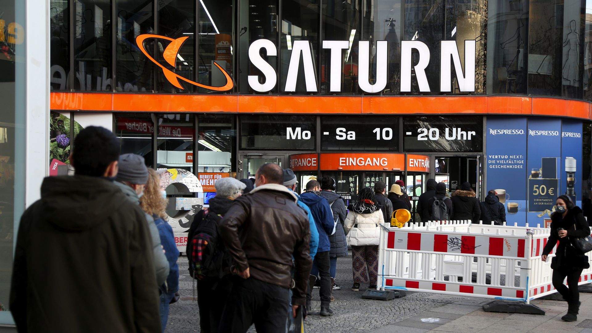 People standing in line outside a Saturn store entrance with orange signage, wearing winter clothes and masks, next to a white construction barrier on a city sidewalk.