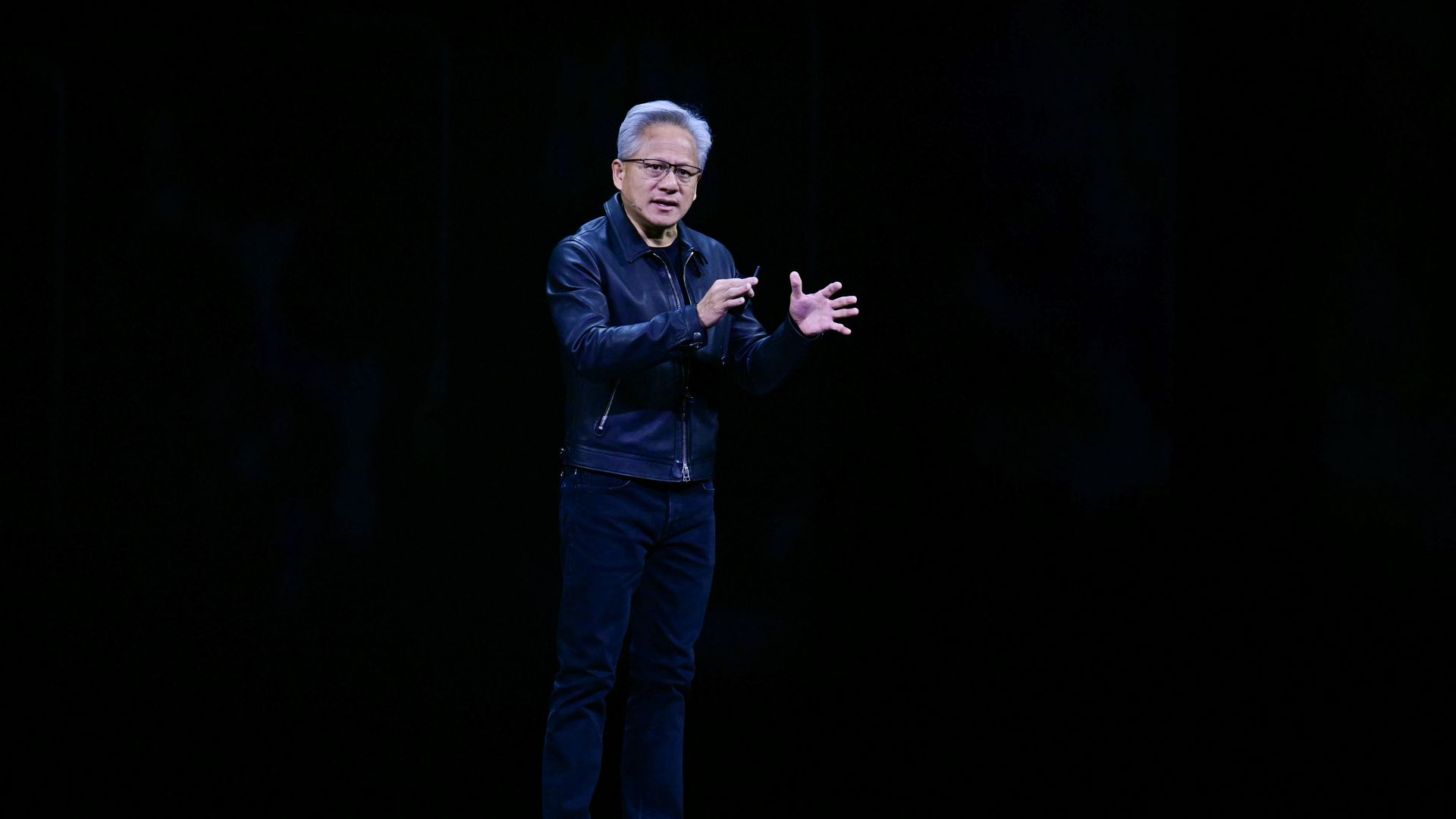 A person in a leather jacket gestures with their hands on a dark stage