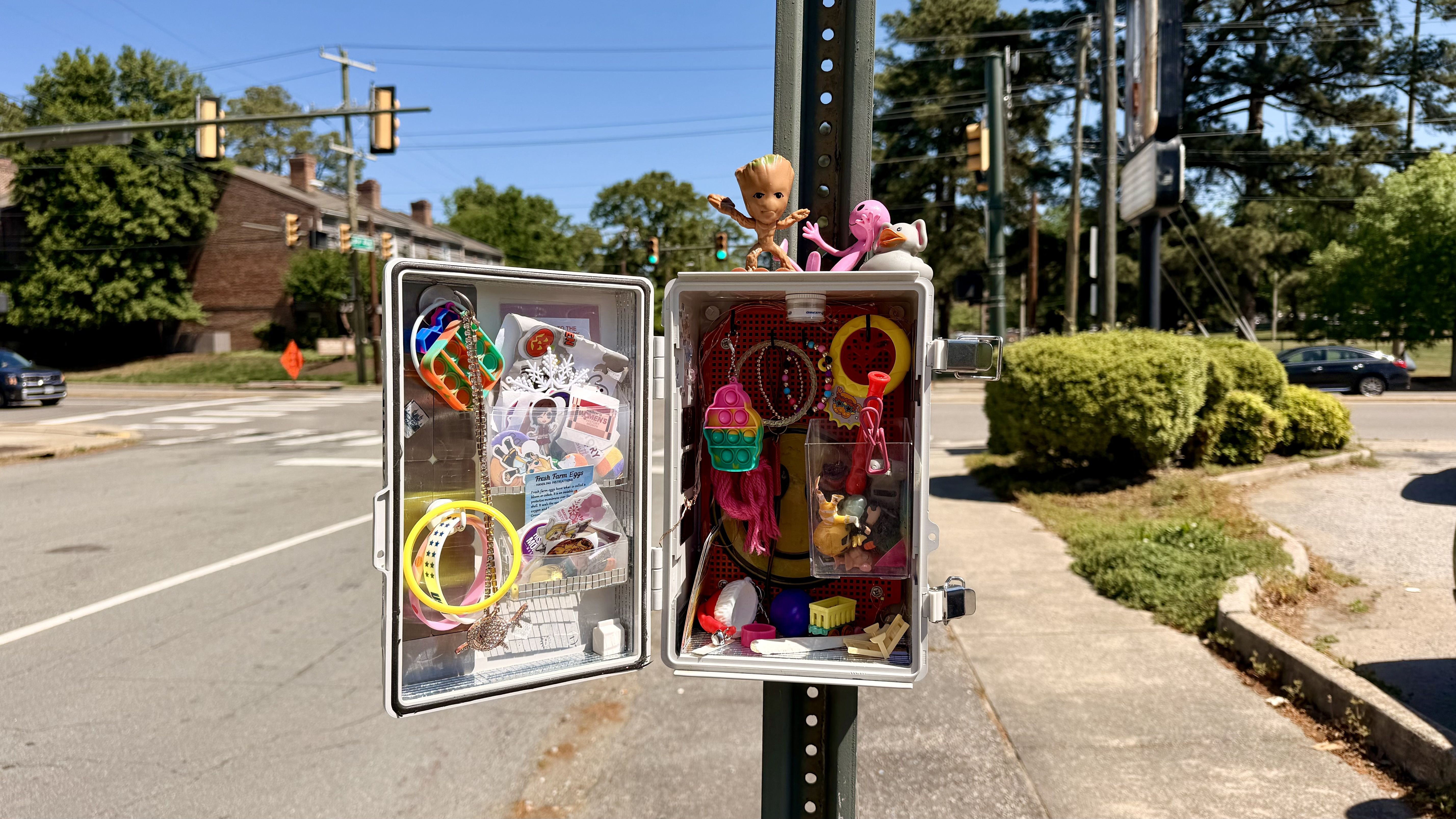 Open two-door toy cabinet on a street pole, packed with colorful toys, beads, and figurines; sunny intersection with traffic lights, cars, and green bushes in the background.