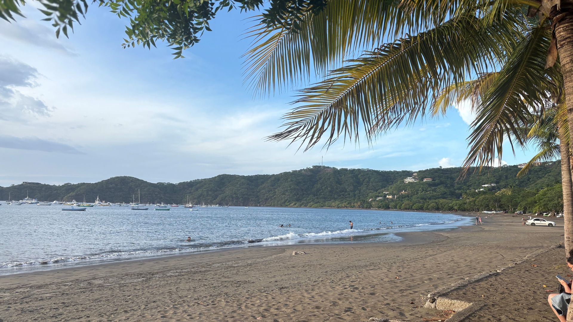 Bright tropical beach with sandy shore, palm fronds arching overhead, calm blue water, distant hills and boats anchored near coastline; a few visitors on shore.