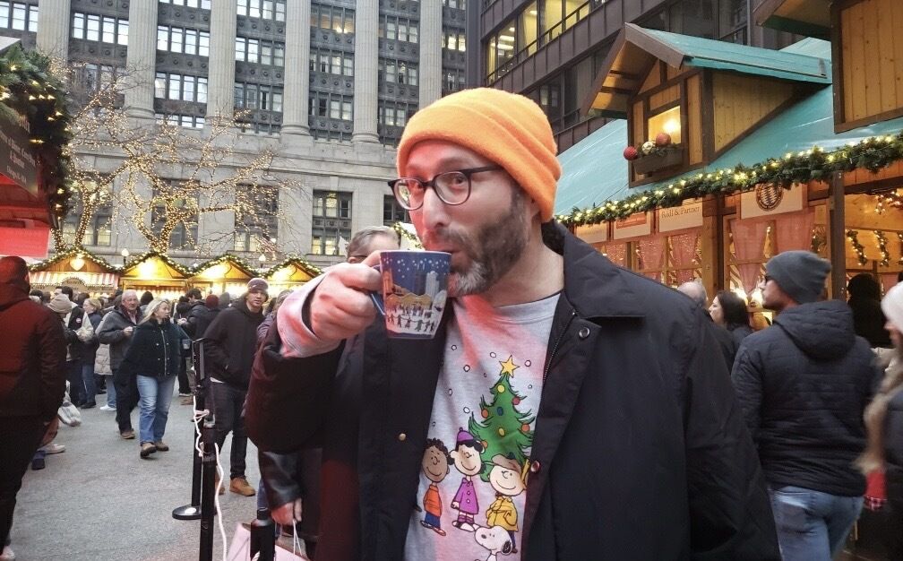 Photo of a man drinking out of a mug at an outdoor market. 
