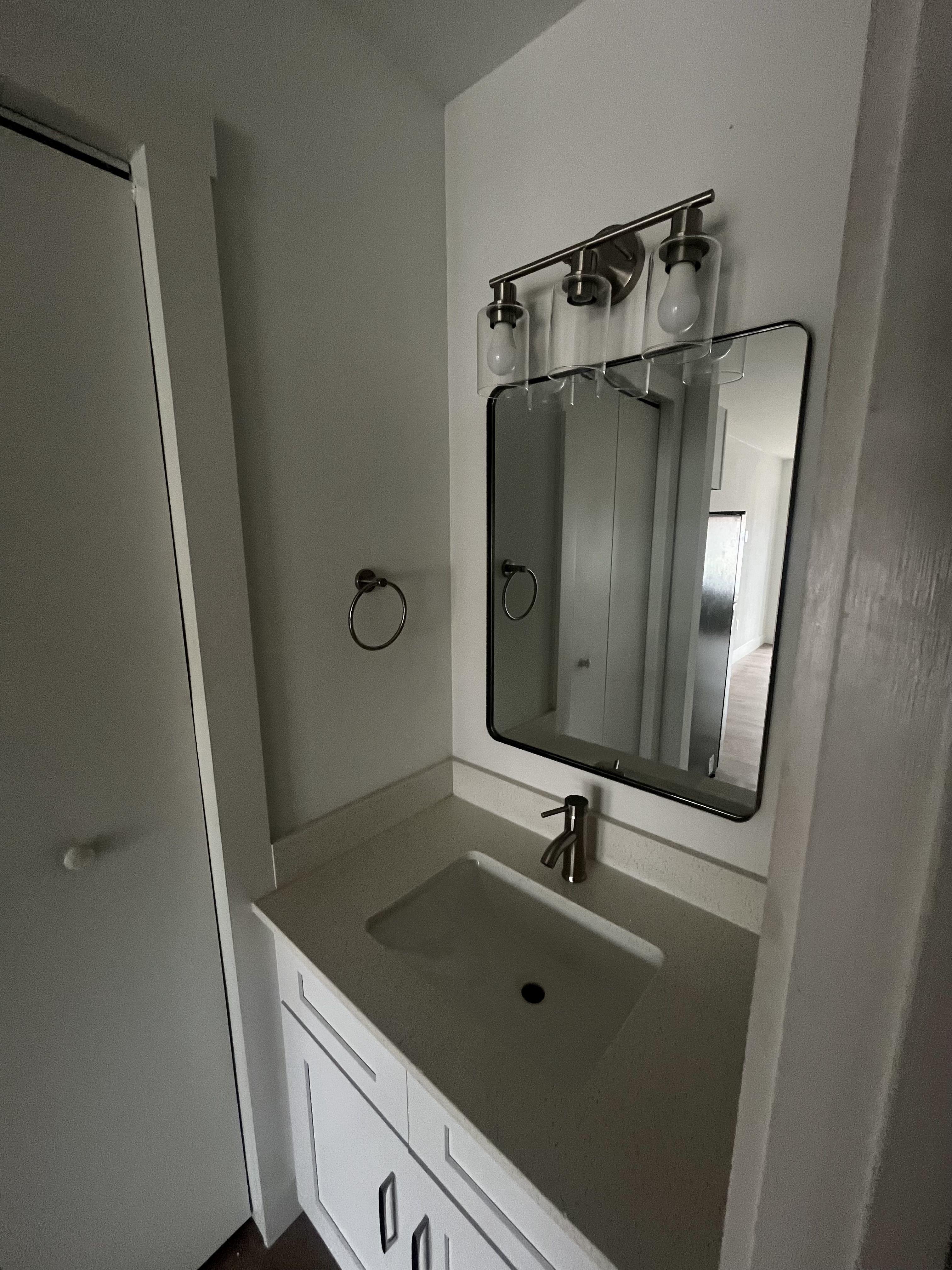 Minimalist bathroom sink area with white countertop and sink, silver faucet, rectangular mirror with black frame, three-bulb light fixture with clear glass shades, and white walls.