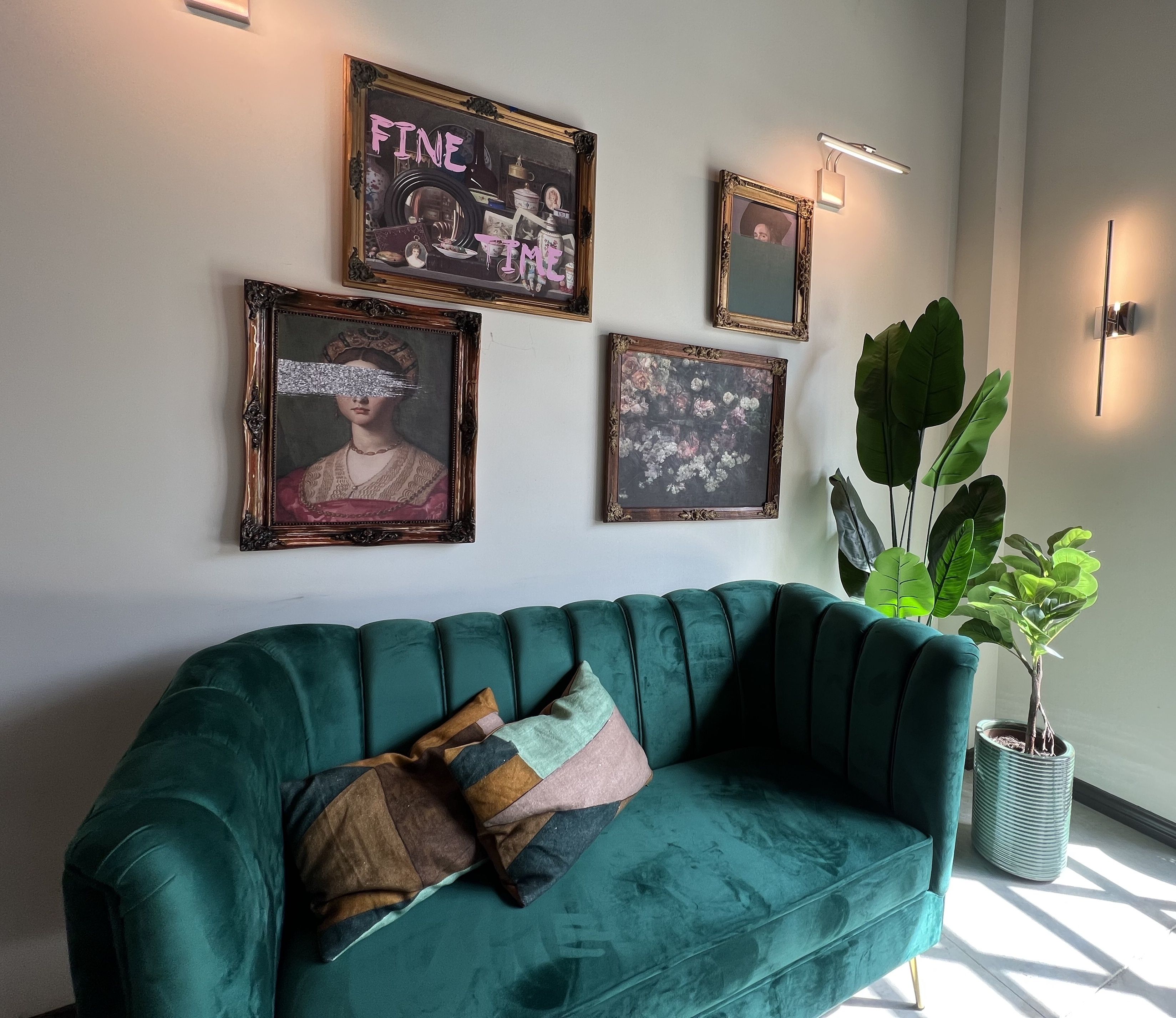 A green velvet couch below art on the wall and by green plants in the corner.