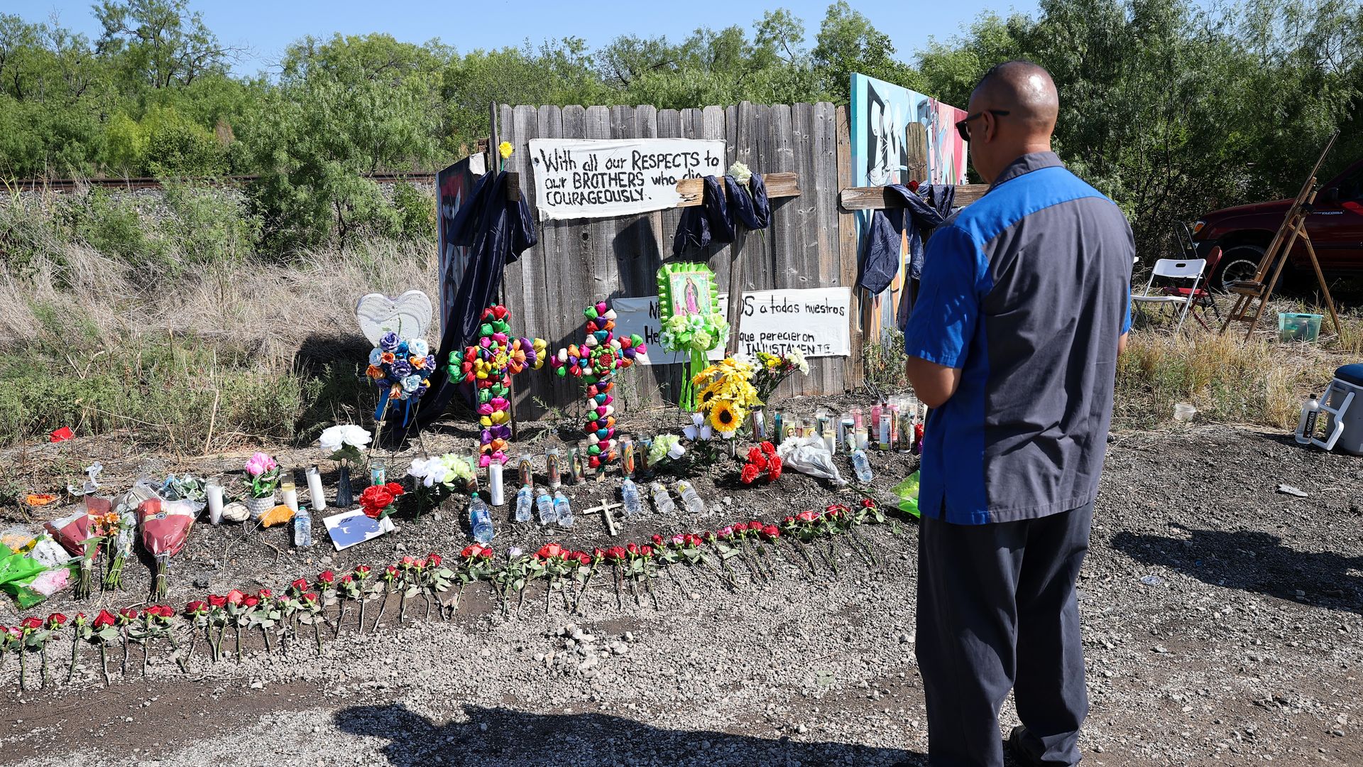 A man stands in front of a makeshift memorial for migrants who died in a smuggling attempt. The memorial includes red roses laid down on the ground, crosses, flowers and signs