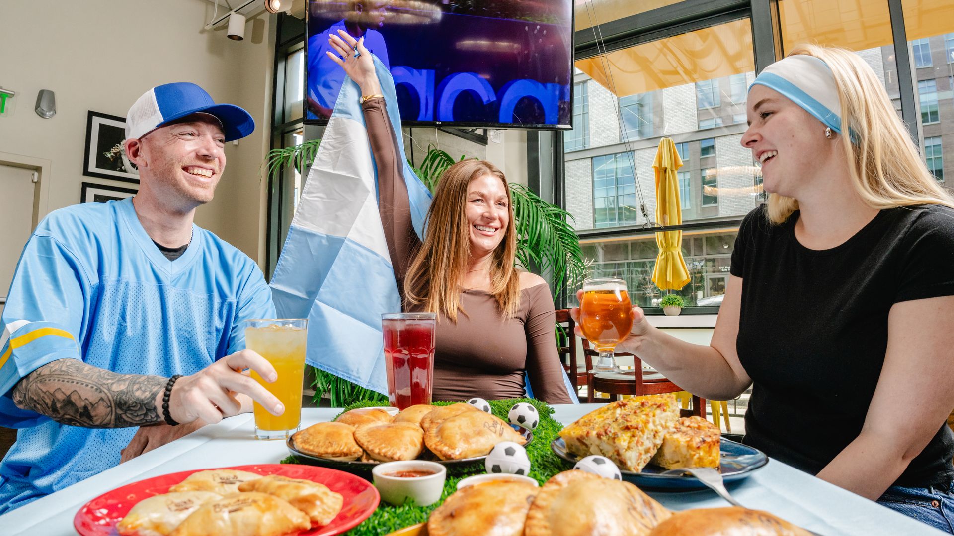 Three people sit while drinking inside a restaurant, with two holding up drinks and one person holding a flag. 