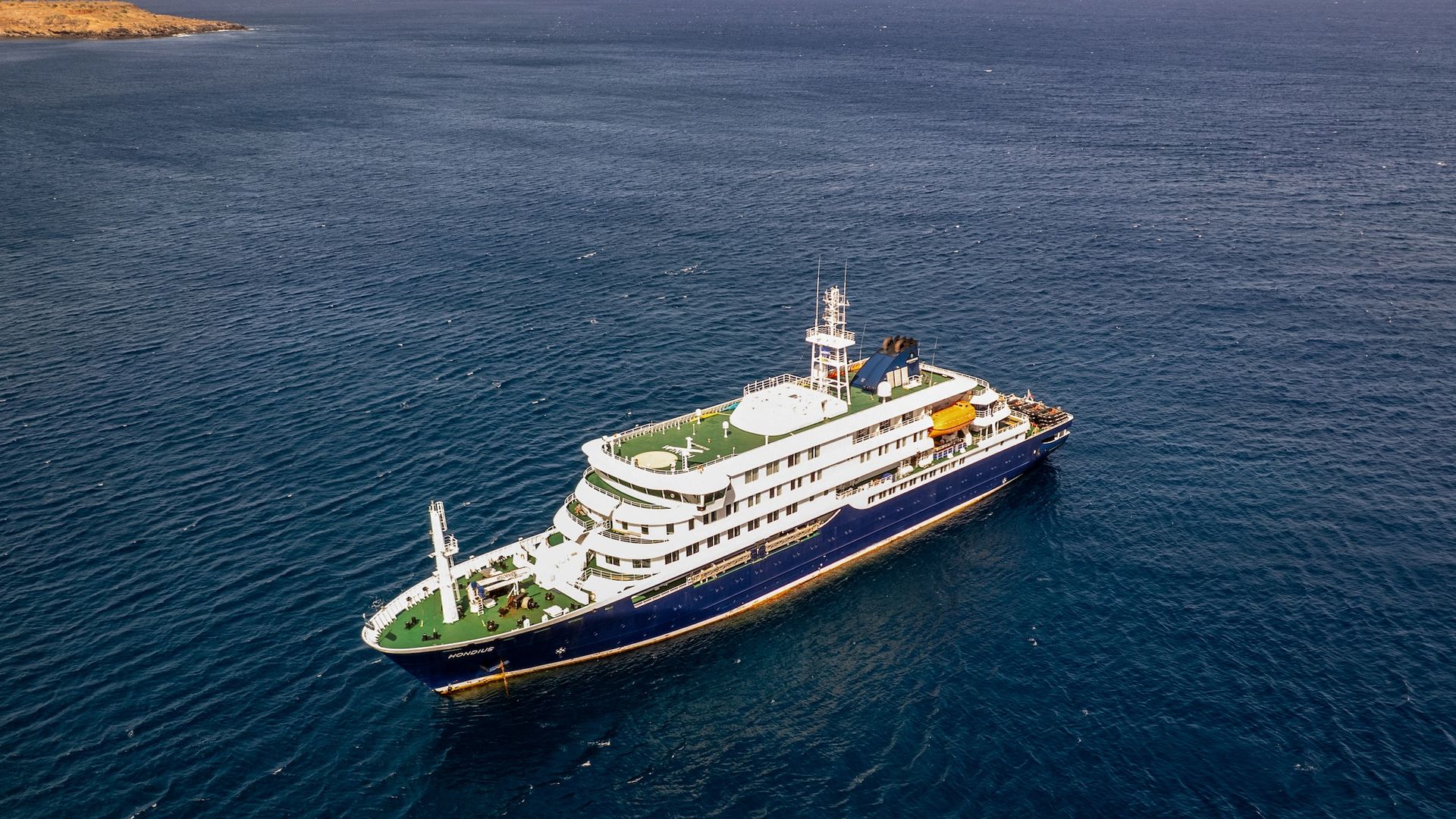 Aerial view of a large white cruise ship with multiple decks and orange lifeboats, sailing through deep blue ocean near a brown rocky coastline on the left and a calm horizon.