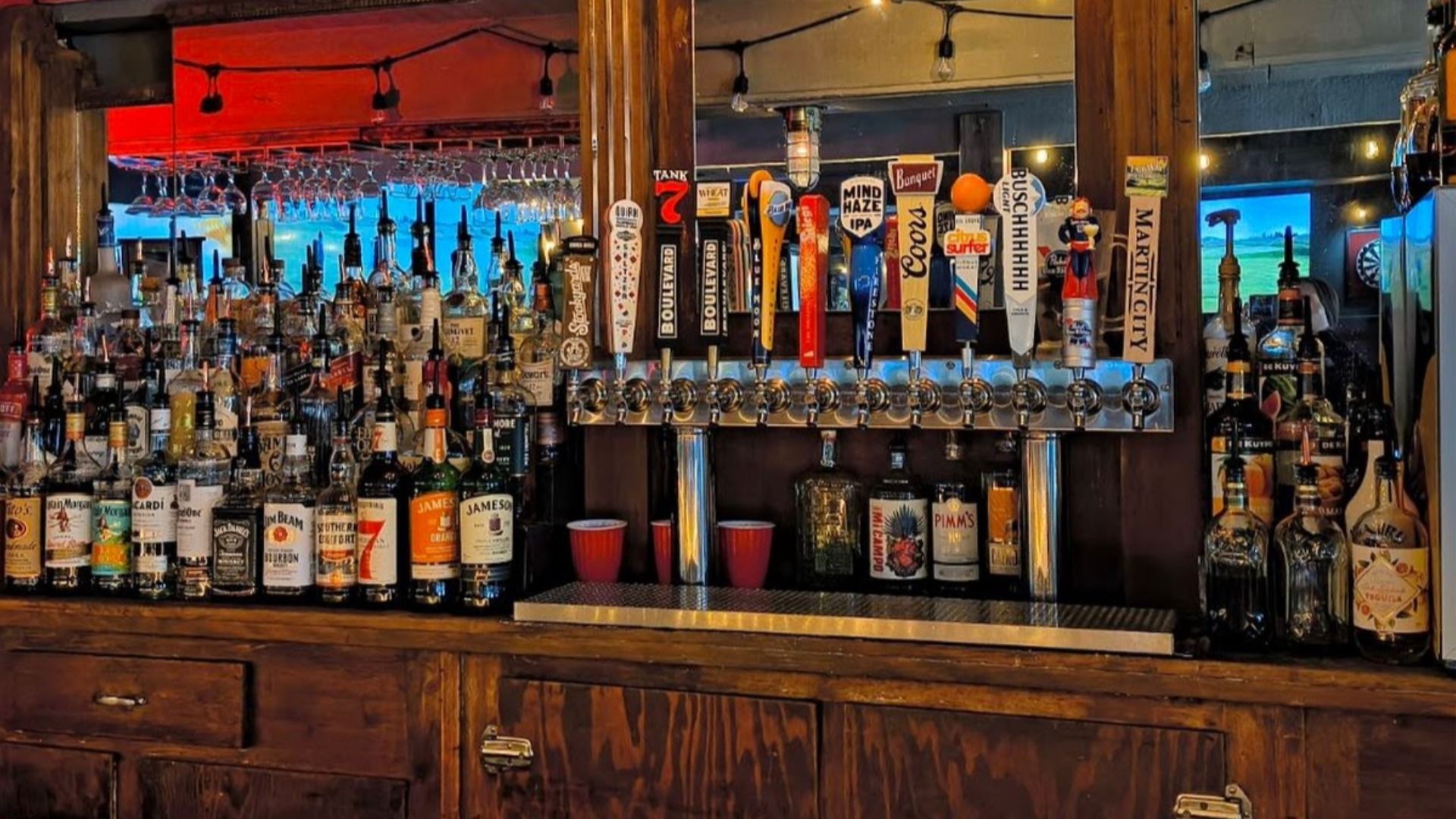 Bar with wooden counters and numerous liquor bottles, beer taps with colorful handles including Coors, Boulevard, and Martin City, and glasses hanging overhead.