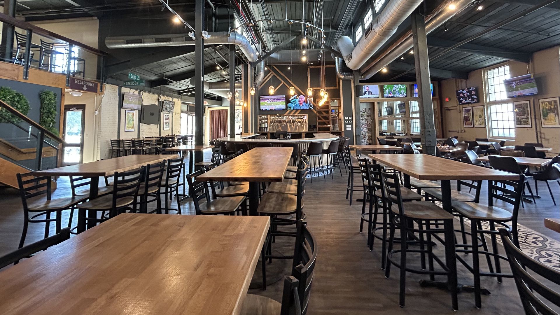 heist brewery. Industrial-style cafe interior with a high ceiling, exposed pipes and ducts, string lights, and numerous wooden tables with black metal chairs; TVs and framed posters along the walls.