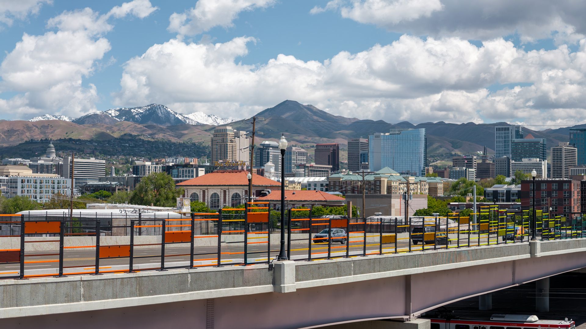 Panoramic cityscape with distant mountains, a highway overpass in the foreground with orange barriers and black fencing, and a mix of glass and brick buildings under a partly cloudy sky.