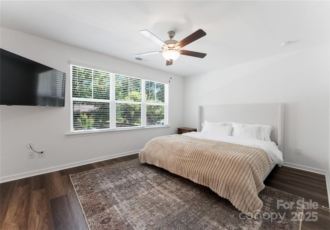 Bright bedroom with white walls, large window with blinds, ceiling fan, king bed with beige striped comforter, white pillows, dark wood nightstand, large patterned rug, and wall-mounted TV.