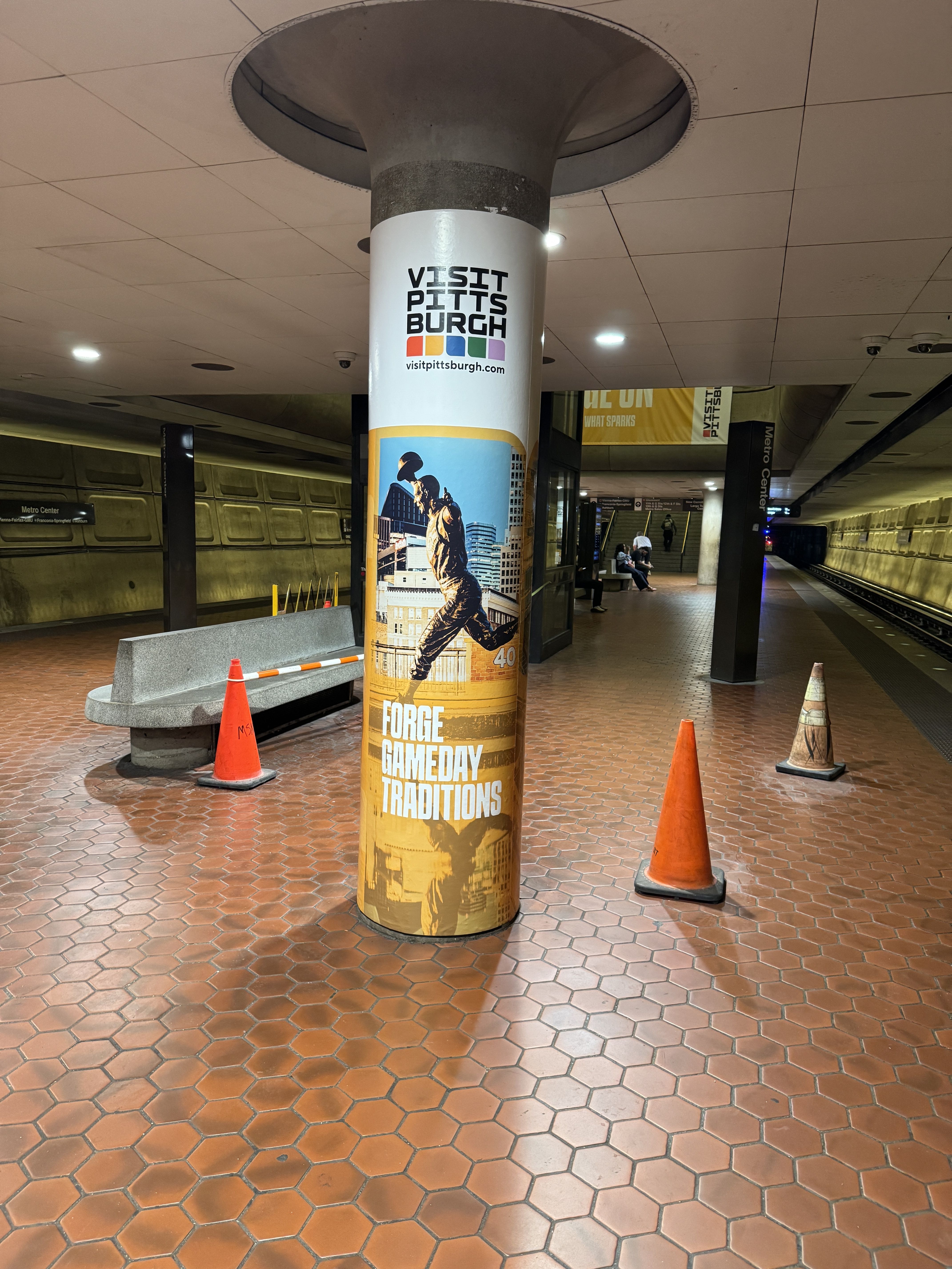 Underground subway pillar wrapped with a "Visit Pittsburgh" poster; colorful squares, orange cones, bench, and brown tile floor, with trains and a few passengers in the background.
