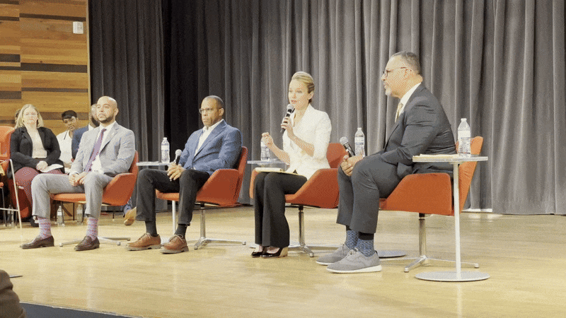 Four panelists sit on stage in red chairs during a discussion; a woman in white holds a microphone speaking, with a gray curtain backdrop and wooden walls on the left.