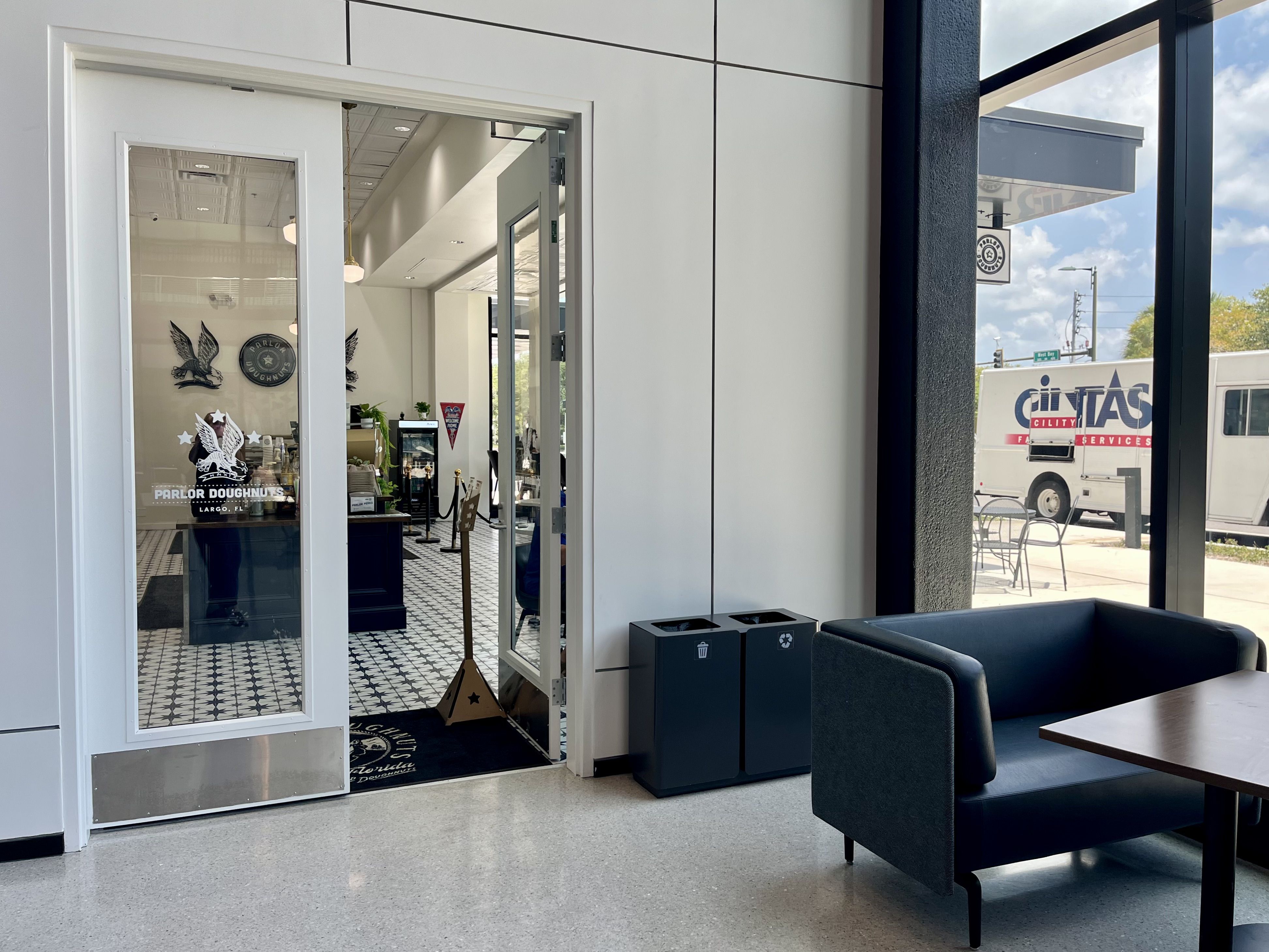 Doorway to a doughnut shop with glass doors; inside features a black-and-white tiled floor, counter, and eagle decals, while outside a Cintas van and blue sky are visible.