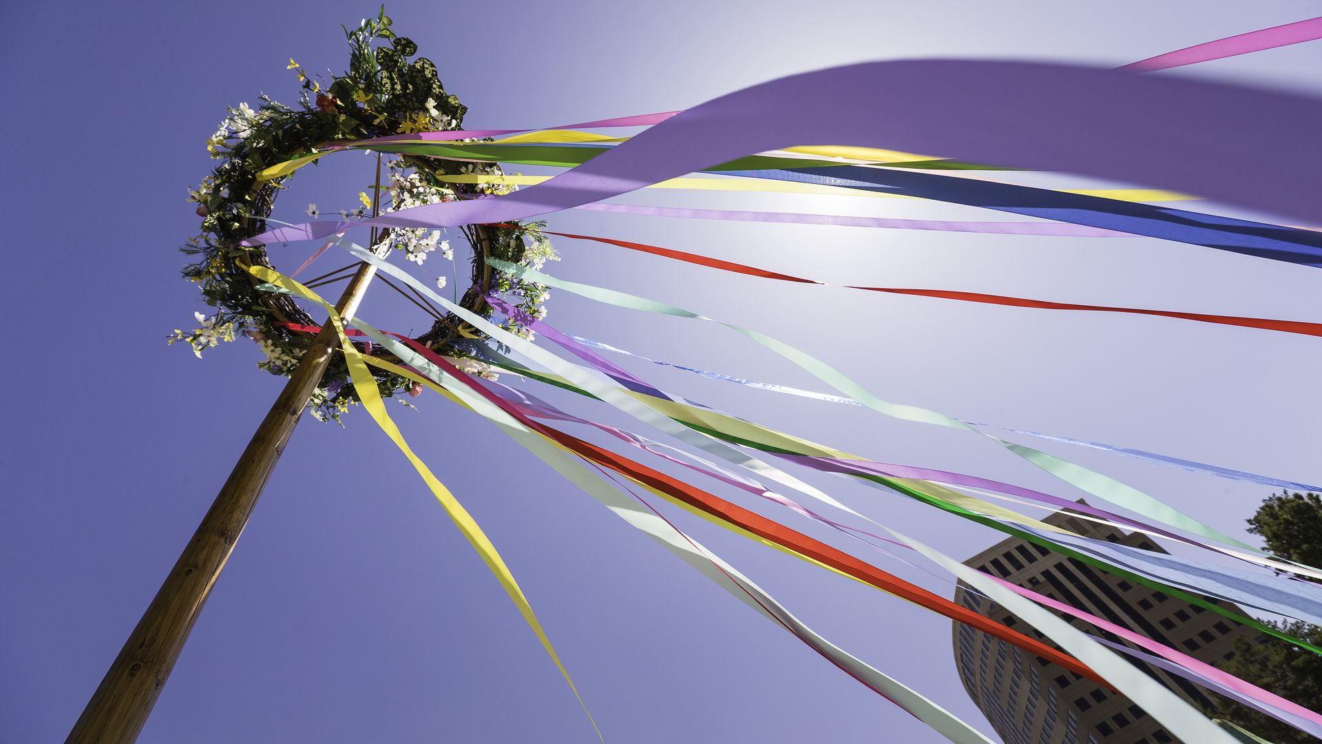 A wooden maypole topped with a flower wreath and long, flowing ribbons in pink, yellow, green, blue, and red fluttering against a clear blue sky; a building is visible in the background.