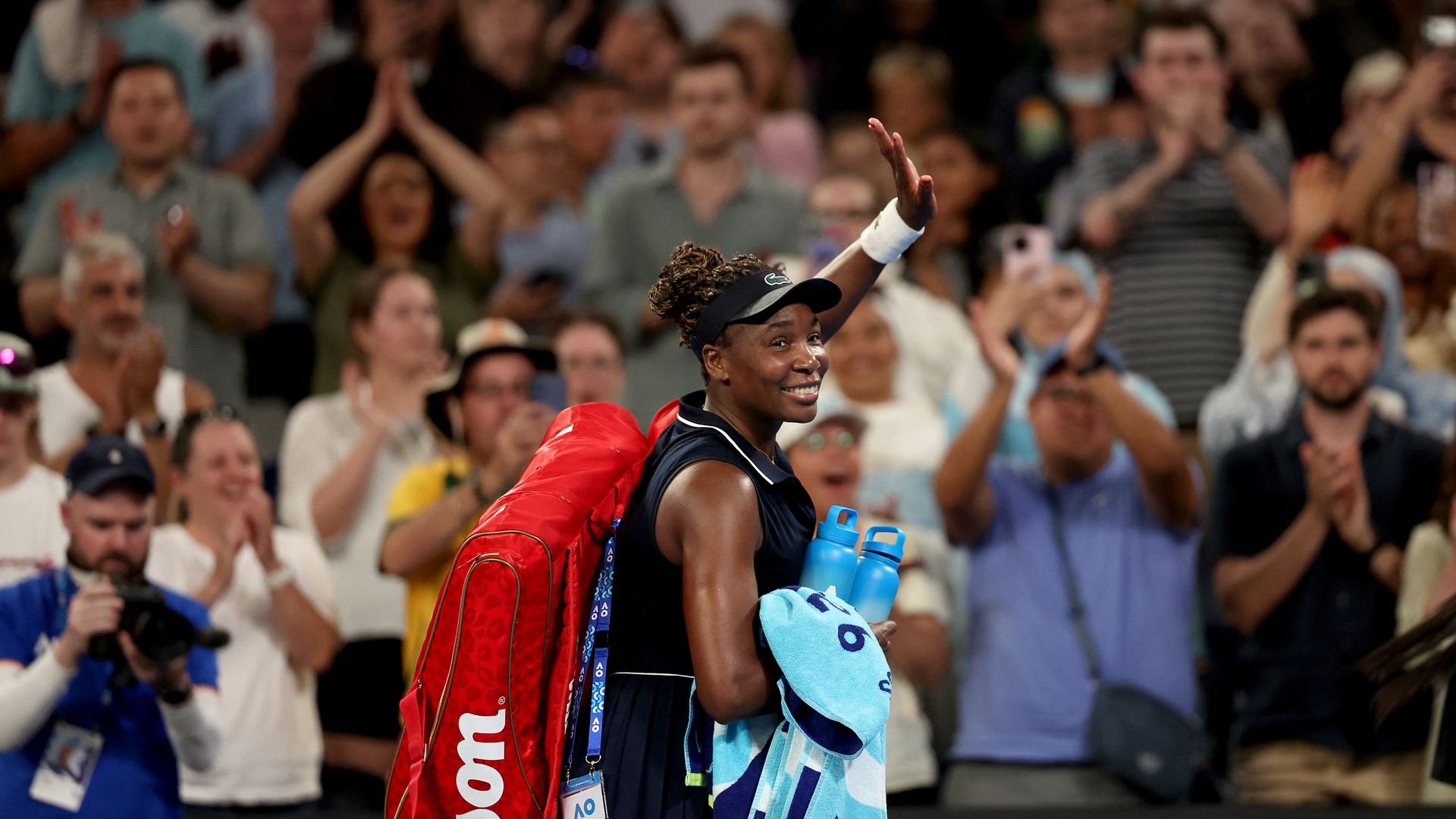 Smiling female tennis player waving to crowd, carrying red Wilson bag, blue towel, and two water bottles, with applauding audience in background.