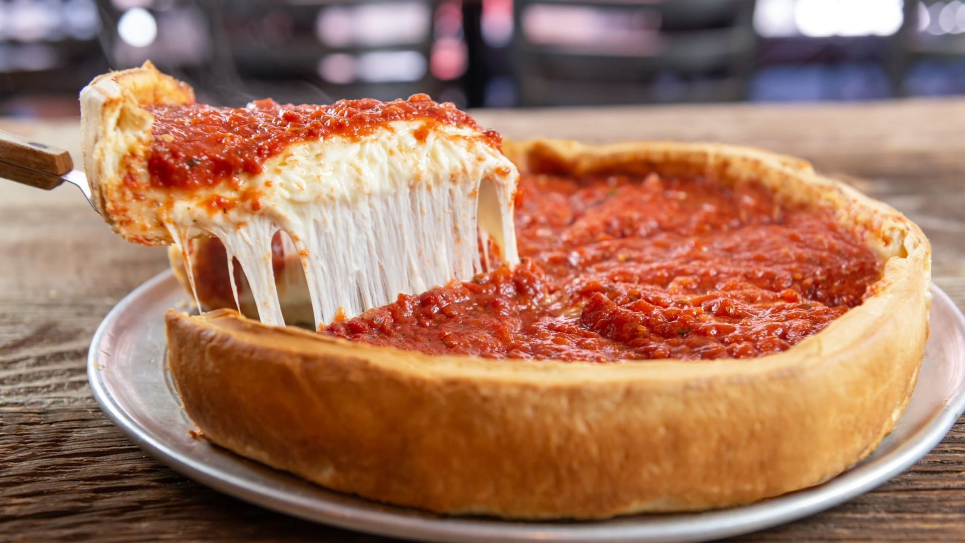 A thick-crust cheese pizza on a silver plate, topped with chunky red tomato sauce; a slice is lifted, long strands of melted mozzarella stretching from the cheese.