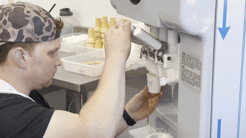 Person with a camouflage cap dispensing white ice cream from a machine into a small paper cup in a kitchen setting with waffle cones in the background.